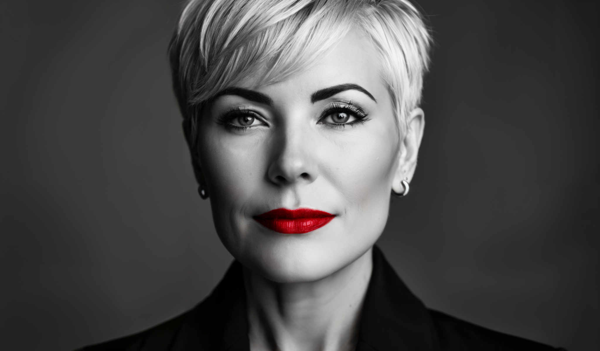 The image is a black and white portrait of a woman with blonde hair. She is wearing red lipstick and a black blazer. Her gaze is directed straight at the camera giving the impression of a serious expression. The background is a plain gray color which contrasts with the woman's attire and accentuates her presence in the image. The absence of color in the photo focuses the viewer's attention on the woman and her expression.