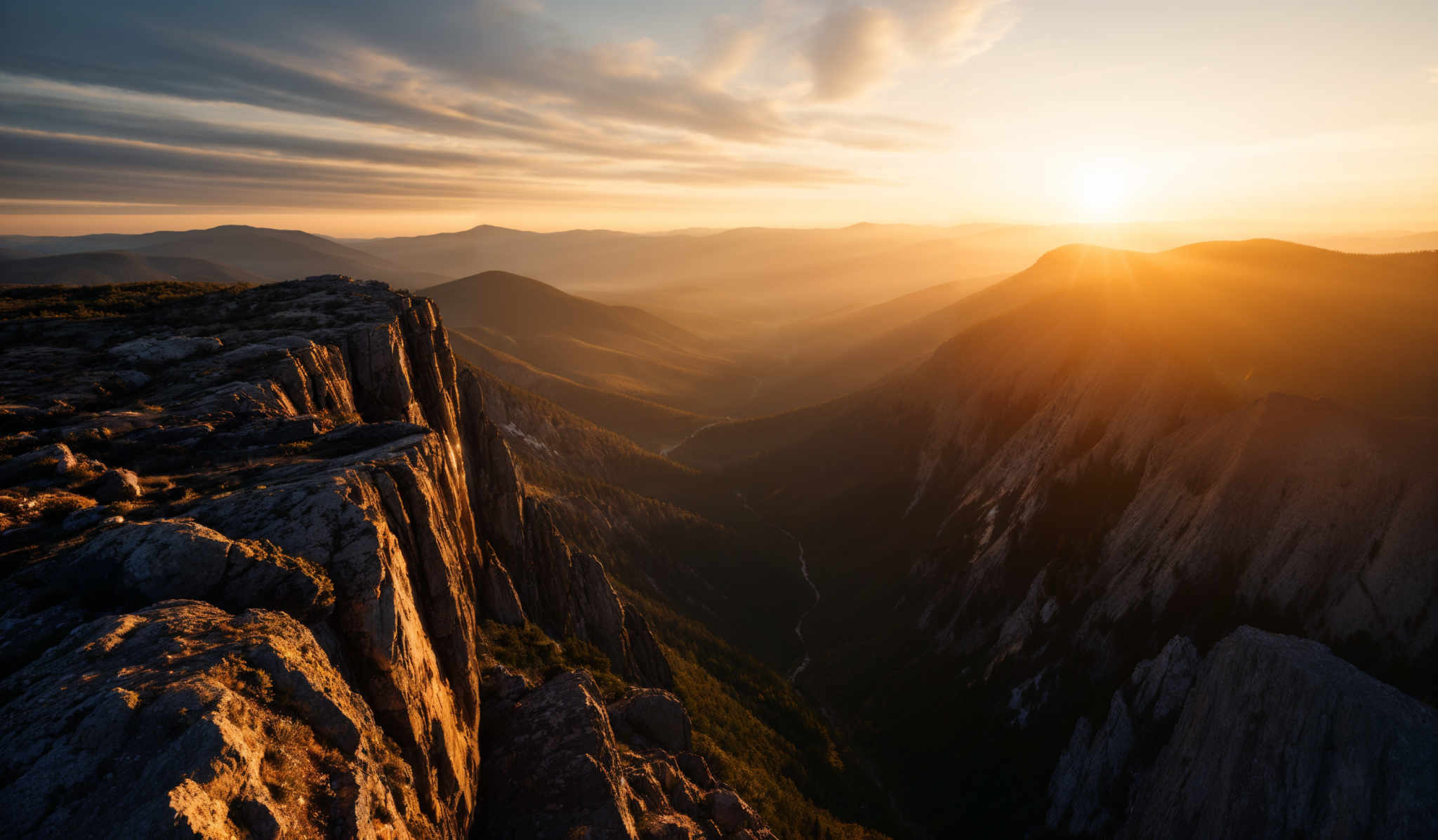 The image showcases a breathtaking landscape during sunset. The sky is painted with hues of orange, gold, and blue, with streaks of clouds illuminated by the setting sun. The sun itself is a radiant golden orb, casting a warm glow over the scene. In the foreground, there's a rugged cliff edge with jagged rocks, some of which are covered in moss or lichen. Below the cliff, a deep valley stretches out, with a winding river or stream visible. The valley is surrounded by mountain ranges, and the distant mountains are bathed in a soft, golden light. The overall scene exudes a sense of tranquility and awe.