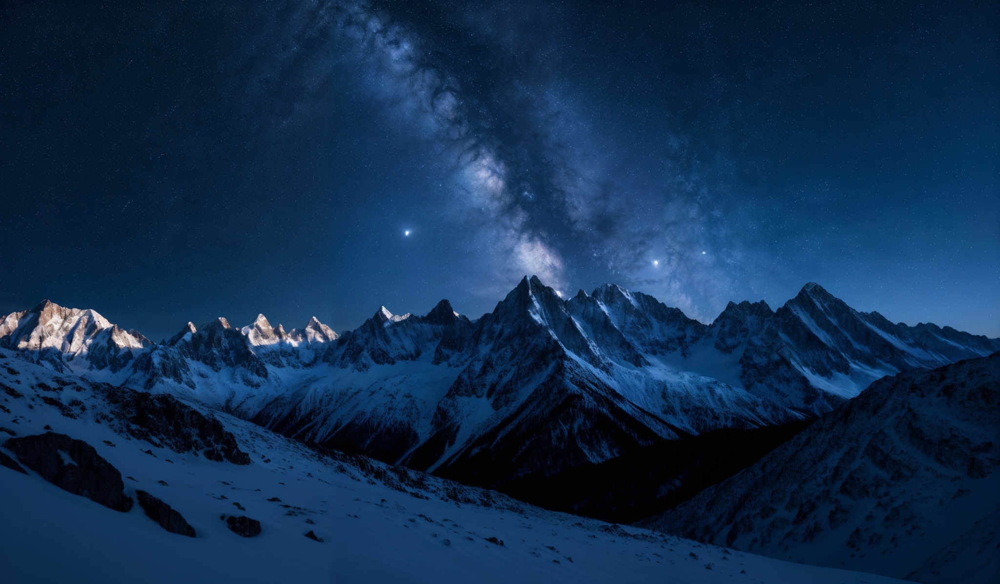 The image showcases a breathtaking night landscape. The dominant colors are deep blues and whites, representing the night sky and the snowy mountains. The Milky Way galaxy, with its dense cluster of stars, stretches across the sky, casting a milky white glow. The jagged peaks of the mountains are covered in snow, and their sharp edges contrast with the smoothness of the sky. The terrain in the foreground is rocky and uneven, with patches of snow scattered around.