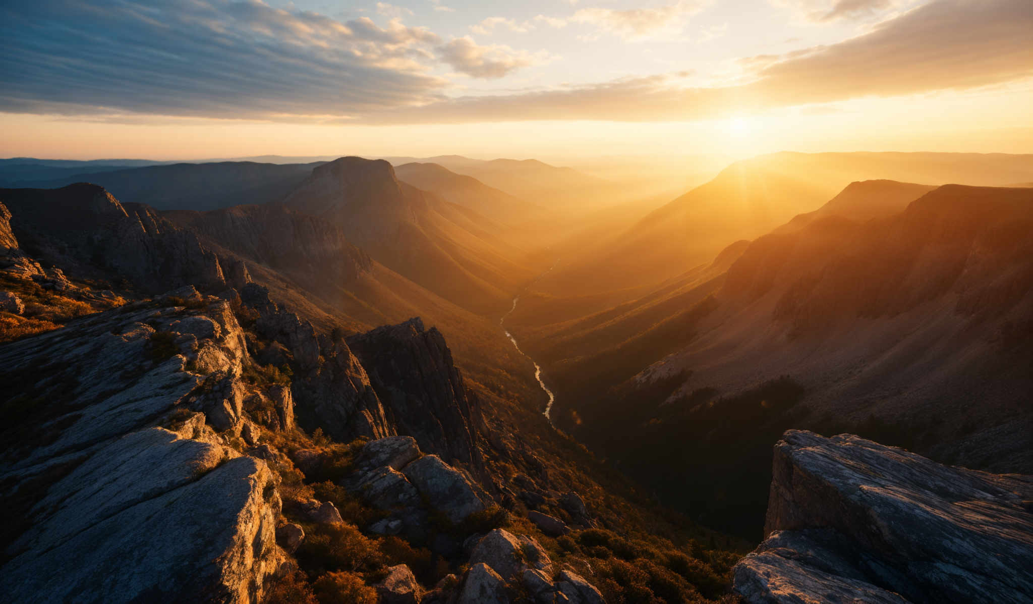 The image showcases a breathtaking landscape during sunset. The sky is painted with hues of blue and orange, with the sun casting a golden glow over the scene. The mountains in the background are rugged and sharp, with deep valleys carved between them. The foreground features rocky outcrops, and there's a winding river or stream visible in the valley below. The play of light and shadow on the mountains adds depth and dimension to the image, making it a captivating view.