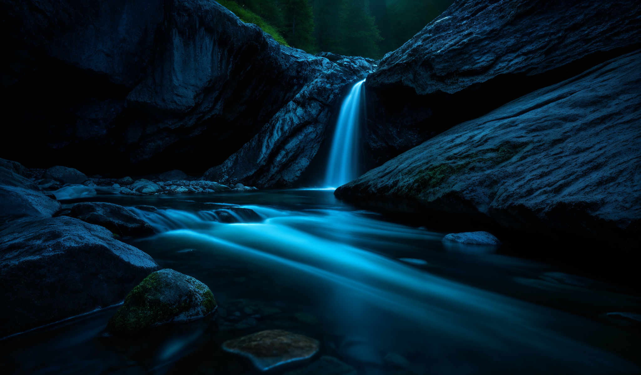 The image showcases a serene natural setting. Dominating the scene are large, jagged rock formations on either side. Between these rocks, a waterfall cascades down, creating a smooth flow of water that contrasts with the roughness of the rocks. The water below the waterfall appears calm, with a silky, bluish hue, possibly due to a long exposure shot. The rocks have varying shades of gray and are interspersed with mossy patches. In the background, there's a hint of a dense forest, adding to the tranquility of the scene.