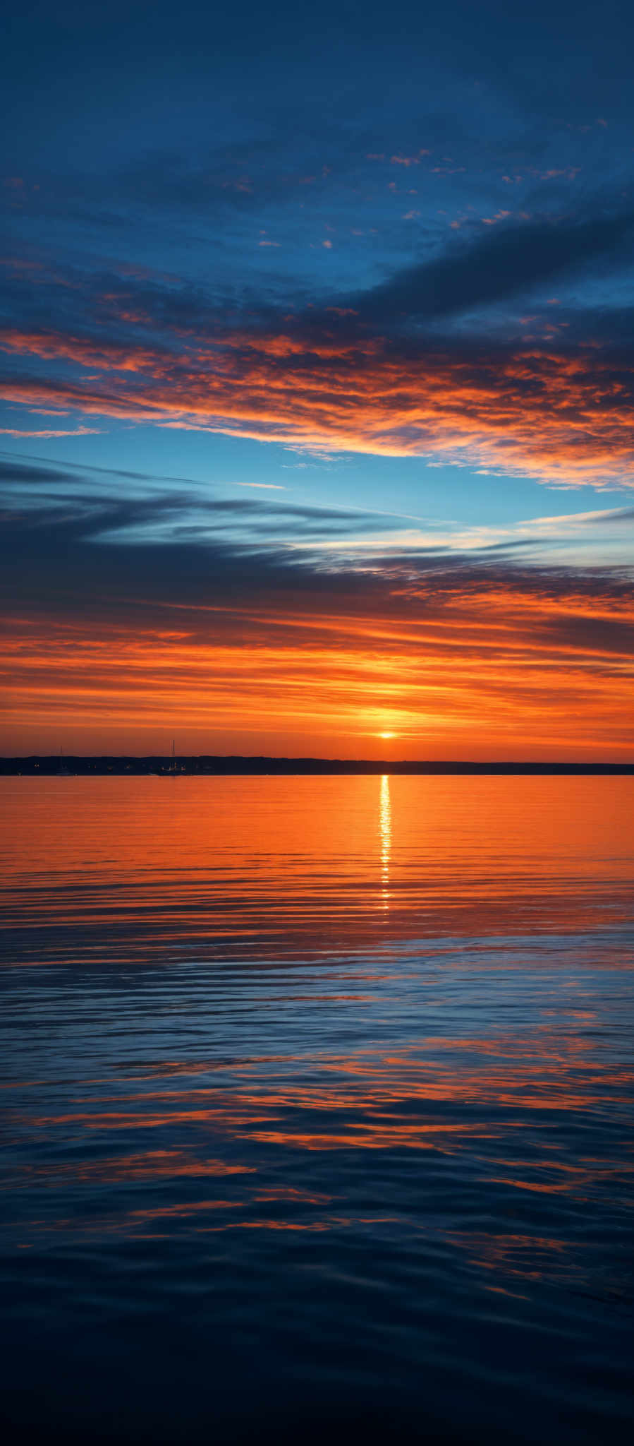 The image showcases a breathtaking sunset over a calm body of water. The sky is painted with a myriad of colors, ranging from deep blues at the top to fiery oranges and reds closer to the horizon. The sun is positioned near the center, casting a brilliant golden hue over the water. Wispy clouds are scattered across the sky, reflecting the sun's colors. The water below mirrors the sky's vibrant colors, creating a harmonious and serene ambiance.