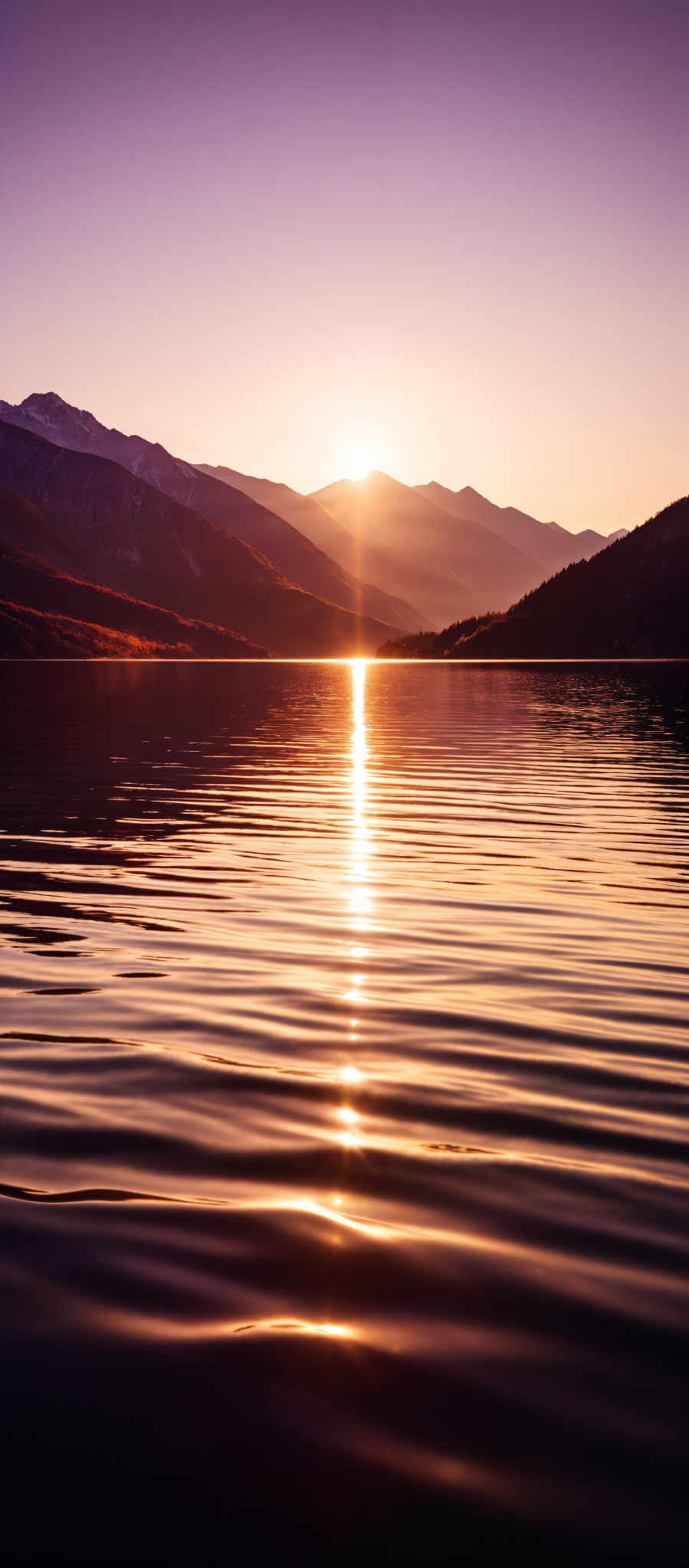 The image showcases a serene landscape during sunset. The sky is painted in hues of purple and pink, transitioning to a golden-orange near the horizon where the sun is setting. The sun casts a radiant glow, creating a reflection on the calm waters of a lake. The lake's surface has gentle ripples, reflecting the sun's light and the surrounding mountains. The mountains, with their rugged terrain, are silhouetted against the sky, adding depth to the scene.