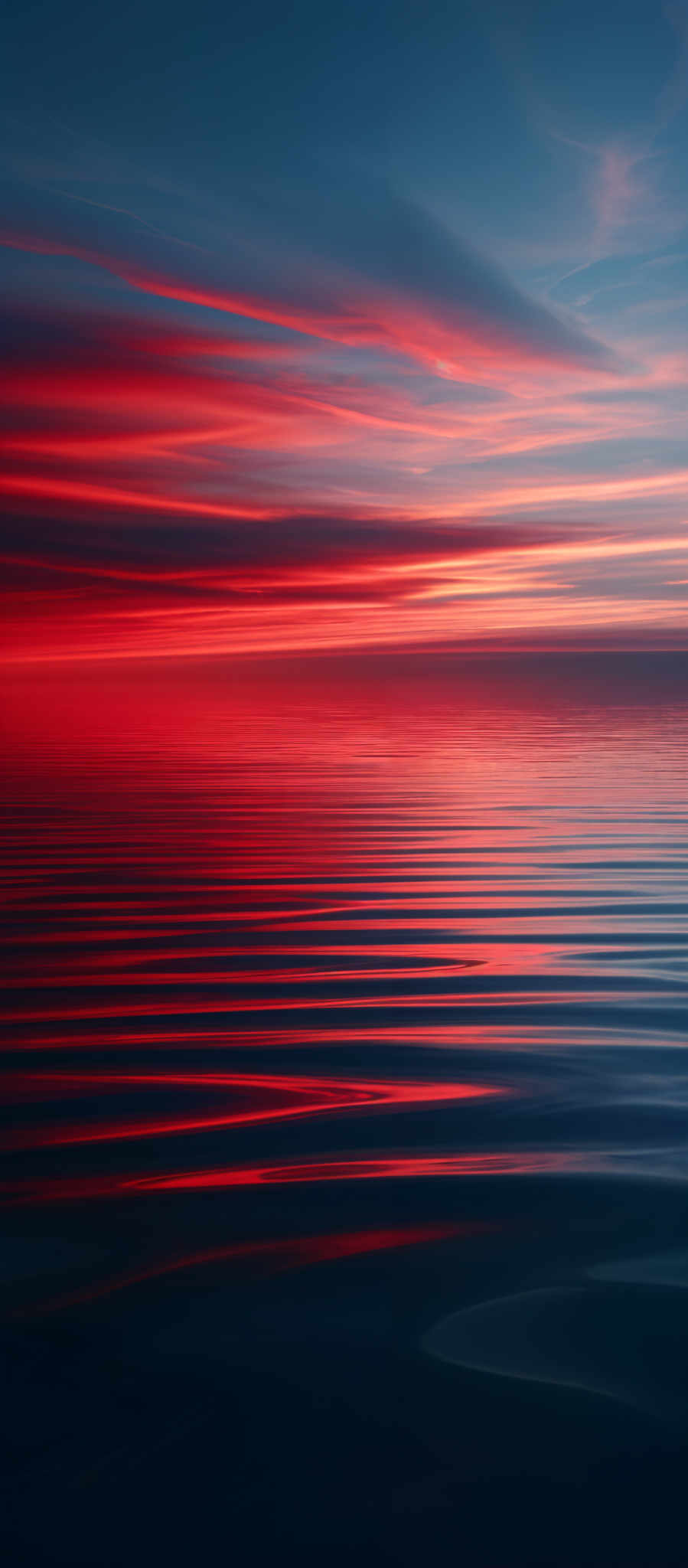 The image showcases a breathtaking sunset over a calm body of water. The sky is painted with hues of deep blue transitioning into fiery reds and oranges. The sun's reflection creates a shimmering pathway of light on the water's surface, with the red and orange tones mirrored perfectly. The horizon is where the sky meets the water, and the gradient of colors creates a serene and mesmerizing effect.