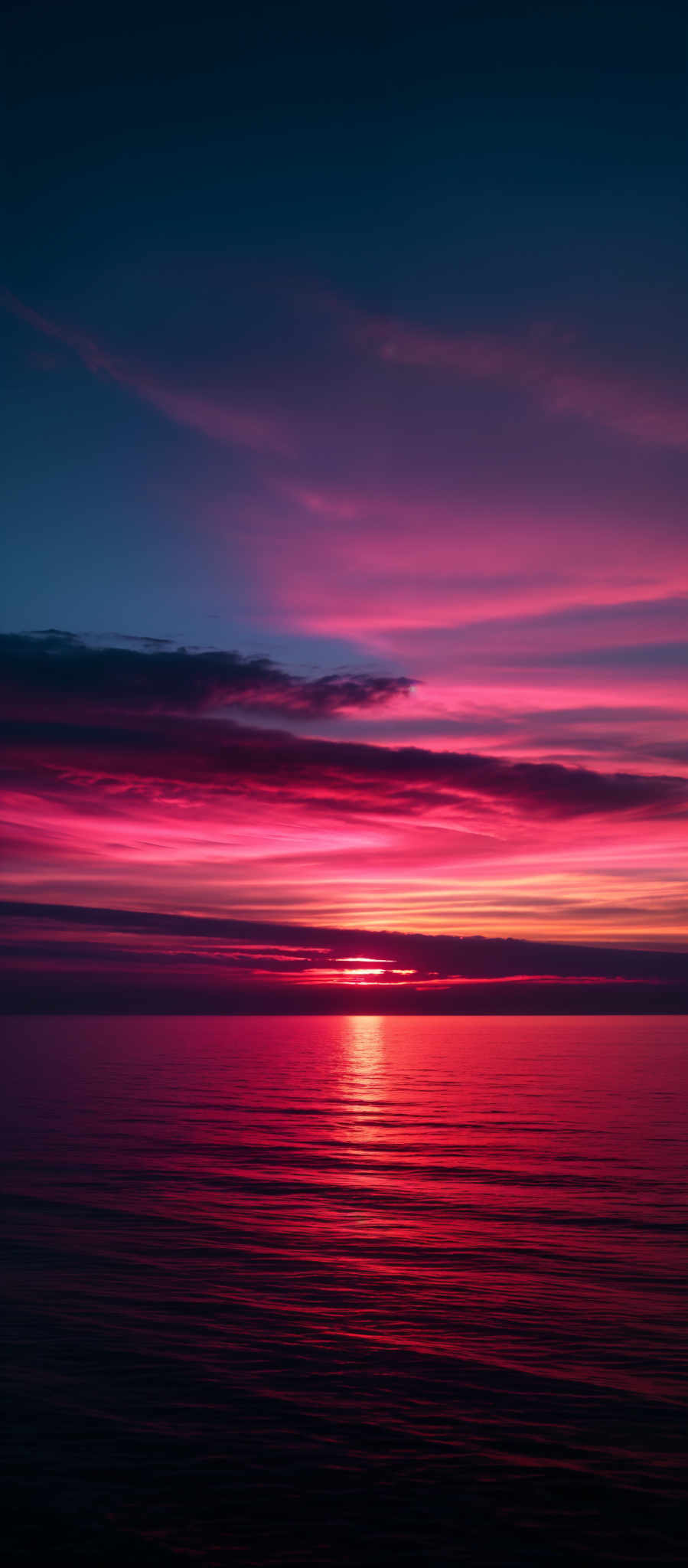 The image showcases a breathtaking sunset over a calm sea. The sky is painted with a gradient of colors, transitioning from deep blue at the top to vibrant shades of pink, orange, and red near the horizon. The sun is partially visible, casting a golden hue over the water. The sea reflects the colors of the sky, creating a mirror-like effect with gentle ripples. The horizon line is clearly visible, separating the sky from the sea.