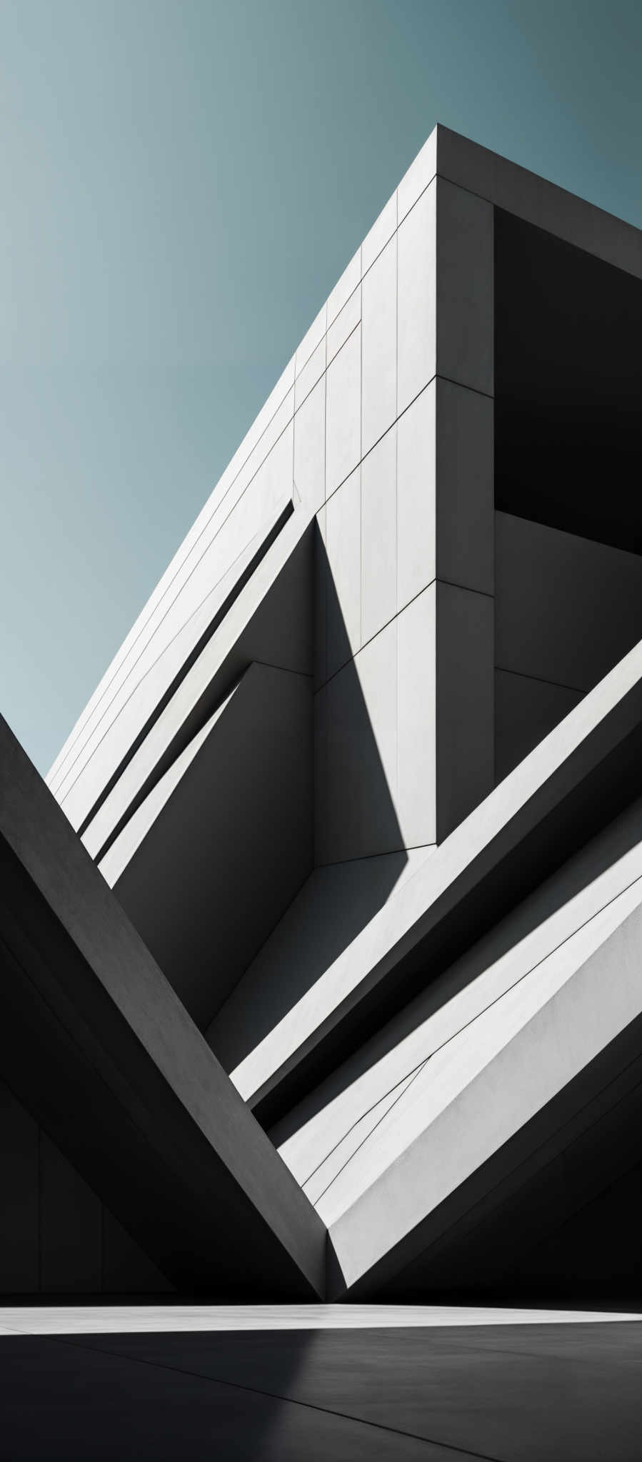 The image showcases a modern architectural structure with sharp geometric shapes. The predominant color is a muted gray, which gives the building a sleek and minimalist appearance. The structure features angular forms, with sharp edges and overlapping planes. The design appears to be made of concrete or a similar material, evident from the smooth and matte finish. The sky in the background is clear, suggesting a sunny day.