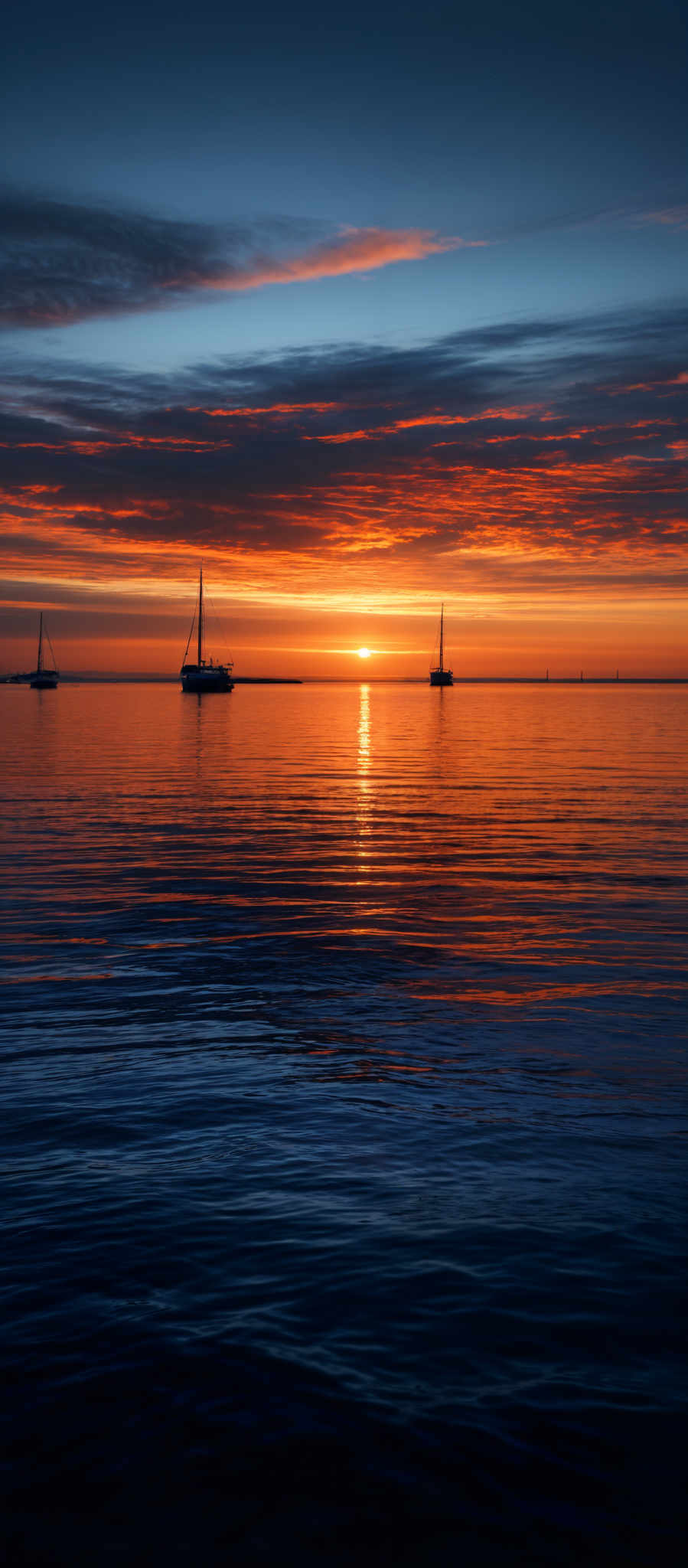 The image showcases a breathtaking sunset over a calm body of water. The sky is painted with hues of orange, gold, and deep blue, with streaks of clouds reflecting the sun's golden light. The sun itself is nearing the horizon, casting a warm glow over the water. On the water, there are three sailboats, each with a tall mast, anchored at a distance from each other. The water's surface is smooth, with gentle ripples reflecting both the sky's colors and the silhouettes of the boats.