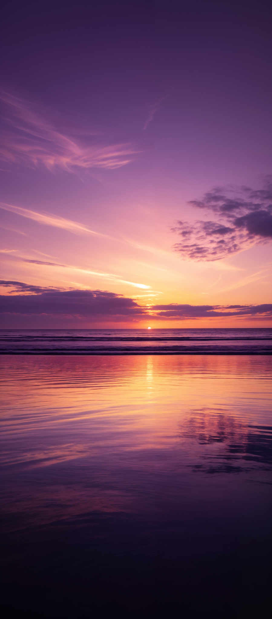 The image showcases a serene sunset over a calm beach. The sky is painted with hues of purple, pink, and orange, with wispy clouds scattered across. The sun is nearing the horizon, casting a golden glow. The beach reflects the colors of the sky, creating a mirror-like effect on the wet sand. The horizon separates the sky from the calm sea, which has gentle waves.