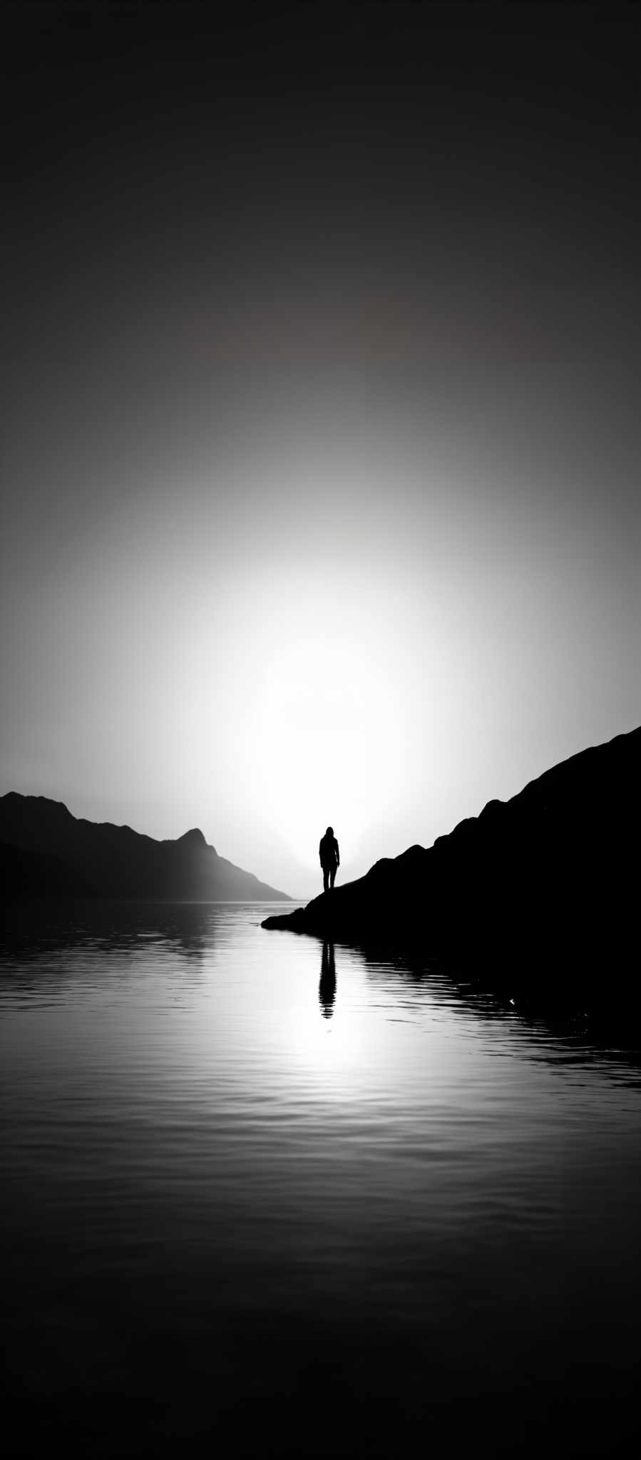 The image is in black and white, showcasing a serene landscape. The sky is vast and expansive, transitioning from a darker shade at the top to a lighter one near the horizon. The horizon reveals a silhouette of mountains. In the foreground, there's a calm body of water reflecting the sky and the silhouettes of the mountains. A solitary figure stands on a rocky outcrop, casting a reflection on the water. The overall mood of the image is tranquil and contemplative.