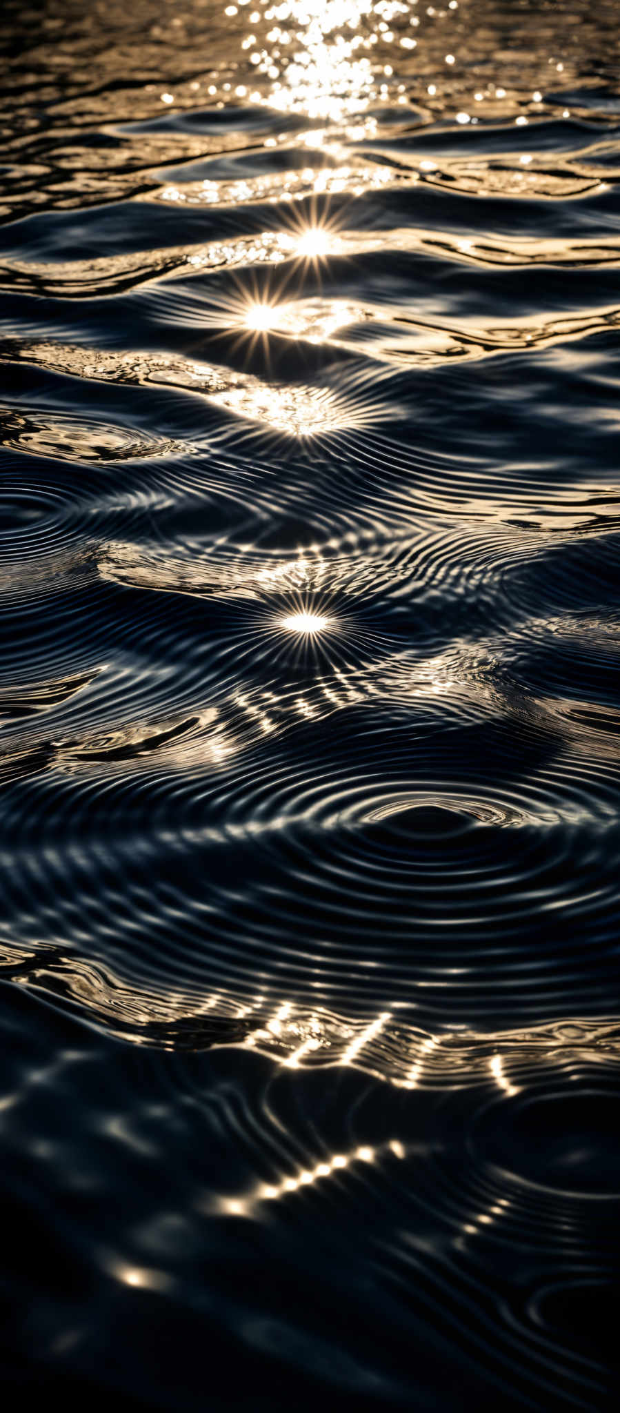 The image showcases a close-up view of water, possibly a lake or a calm sea. The water is dark, almost black, with shimmering golden hues reflecting the sunlight. There are concentric circles on the water's surface, likely created by ripples. The sunlight creates a radiant effect, with bright spots and starbursts scattered across the water.