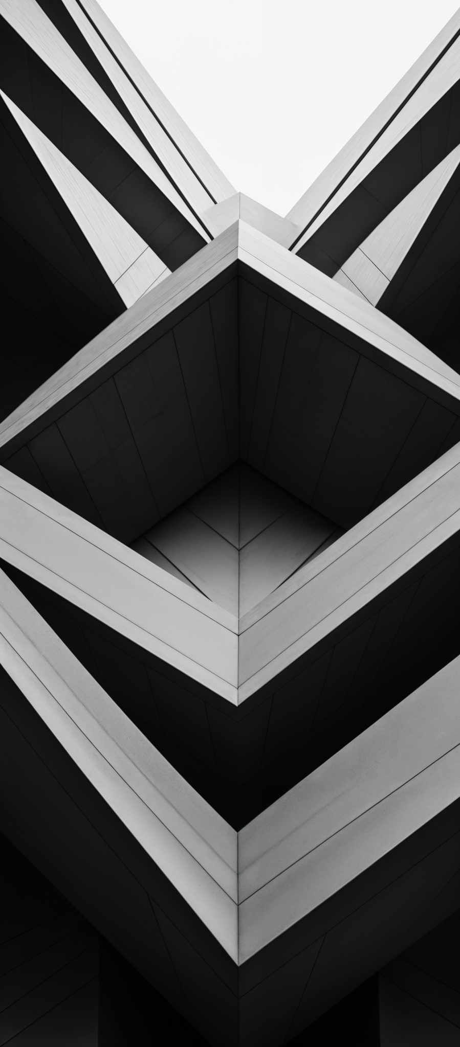 The image showcases a monochromatic architectural design. It features sharp, angular structures made of what appears to be concrete or a similar material. The design is symmetrical, with repeating patterns of interlocking triangles and rectangles. The shapes are predominantly dark, contrasting with the lighter background, creating a visually striking effect.