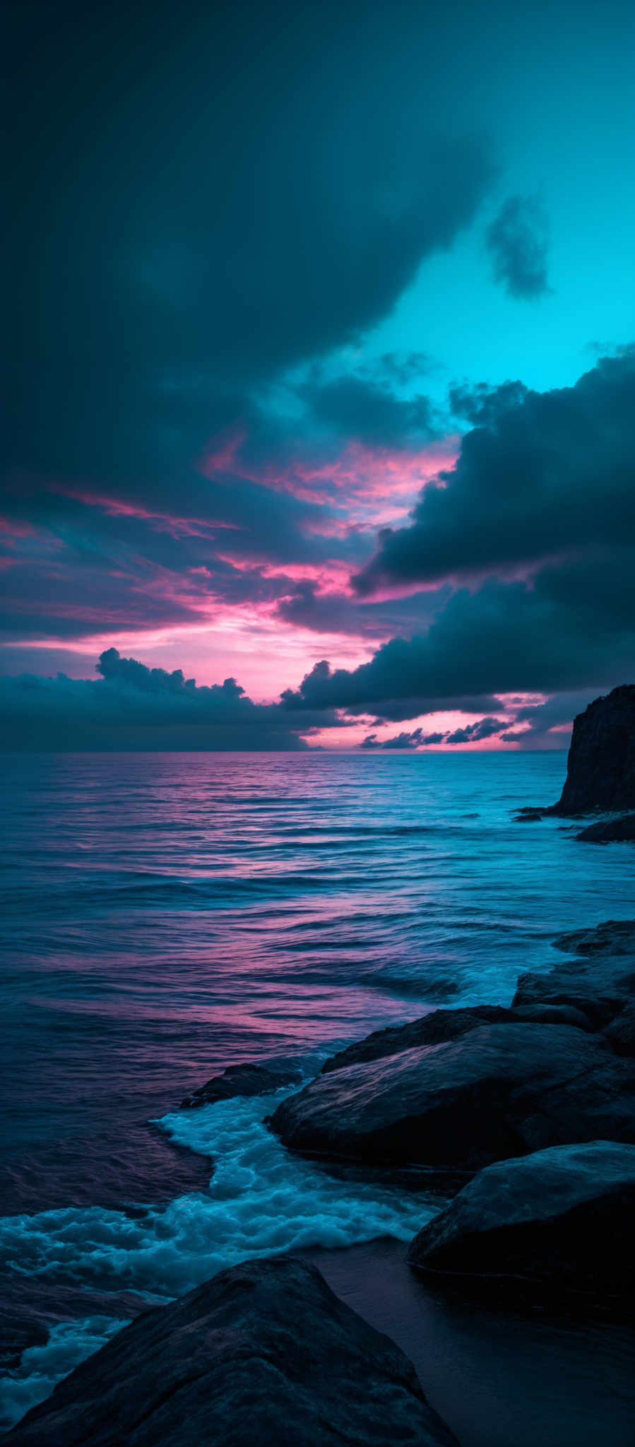 The image showcases a breathtaking seascape during what appears to be either dawn or dusk. The sky is painted with hues of deep blue, purple, and pink, with dark clouds scattered across. The horizon reveals the calm sea, reflecting the colors of the sky. On the right side, there's a rugged cliff with some vegetation, and the waves gently crash against the rocks. The overall mood of the image is serene and contemplative.