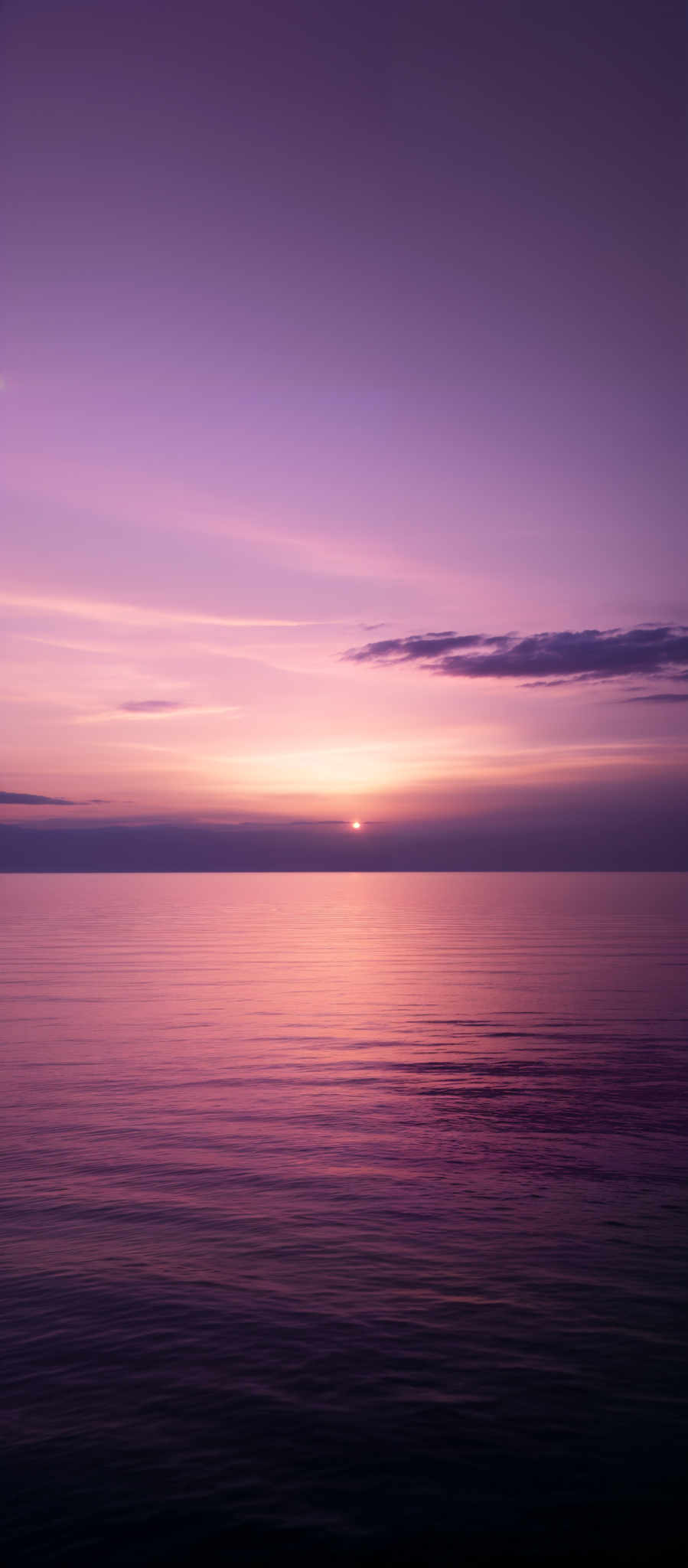 The image showcases a serene sunset over a calm body of water. The sky is painted in hues of purple, pink, and orange, with the sun setting just above the horizon. The water reflects these colors, creating a gradient of purples and pinks. There are also some clouds visible, adding depth and texture to the sky.