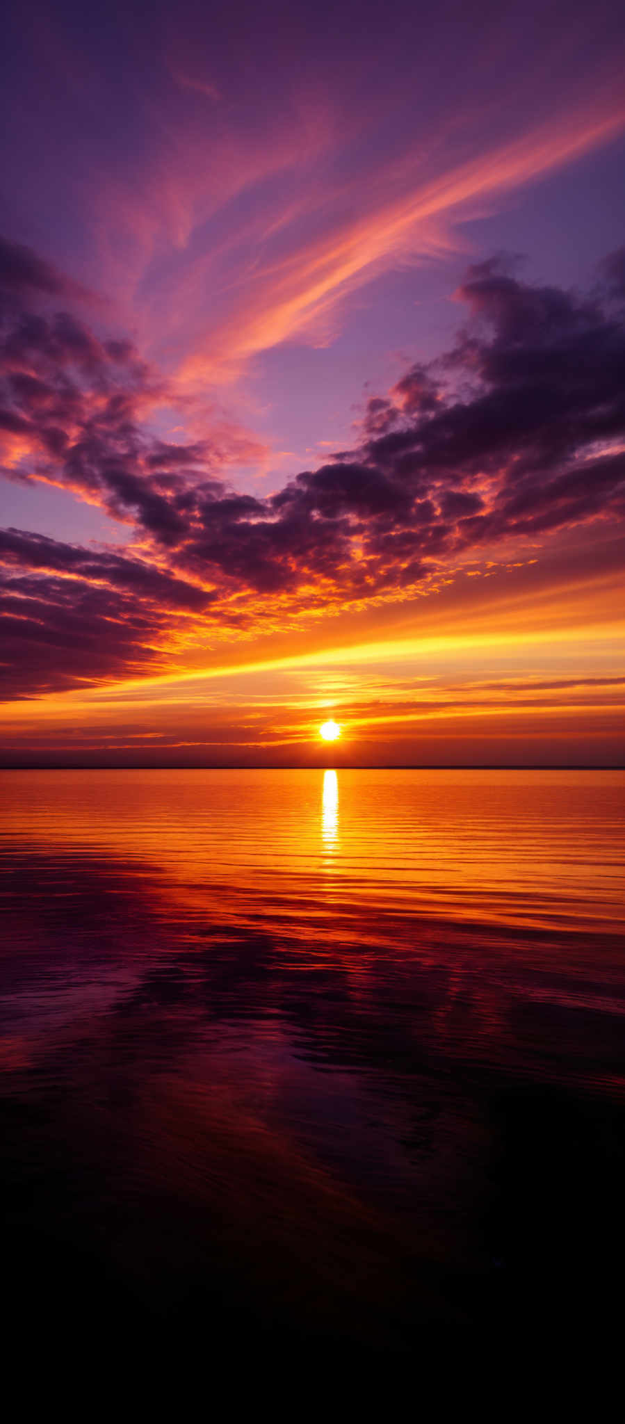 The image showcases a breathtaking sunset over a calm body of water. The sky is painted with a myriad of colors, ranging from deep purples and blues at the top to fiery oranges and yellows near the horizon. The sun is positioned just above the water, casting a brilliant reflection on the water's surface. The clouds are scattered across the sky, with some appearing wispy and others more dense, adding depth and texture to the scene.