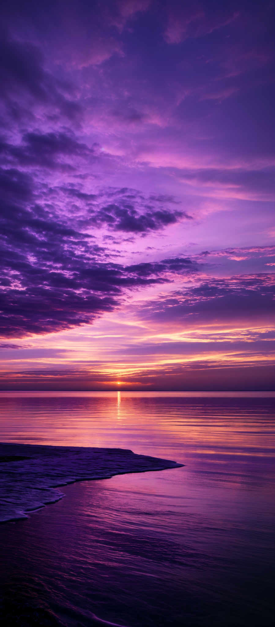 The image showcases a breathtaking sunset over a calm sea. The sky is painted with a myriad of colors, ranging from deep purples and blues to fiery oranges and yellows. The clouds are scattered across the sky, reflecting the hues of the sunset. The sun itself is nearing the horizon, casting a golden glow over the water. The sea reflects these colors, creating a mirror-like effect. On the left side, there's a shoreline with gentle waves lapping at the beach.