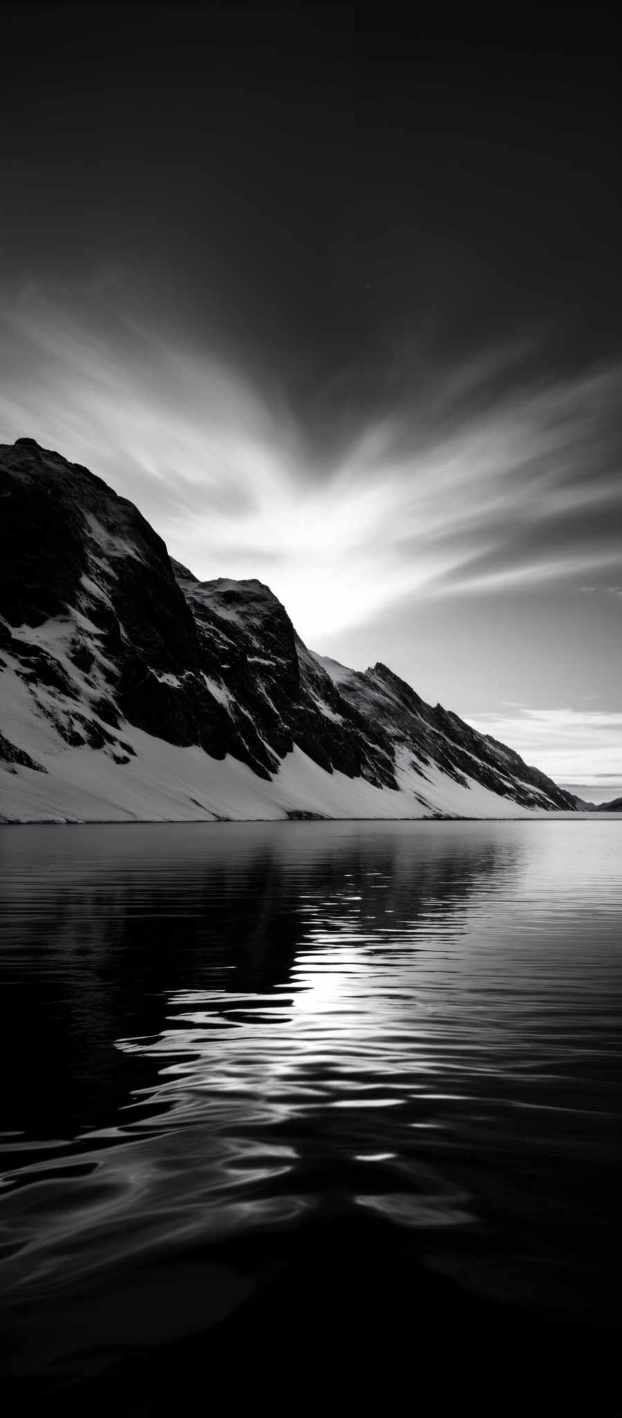 The image is in black and white, showcasing a serene landscape. The dominant shape is that of a mountain range with jagged peaks, partially covered in snow. The mountains are reflected in a calm body of water, creating a mirrored effect. Above, the sky is filled with streaks of clouds, possibly indicating a long exposure shot, which gives the clouds a smooth, flowing appearance. The overall mood of the image is tranquil and contemplative.