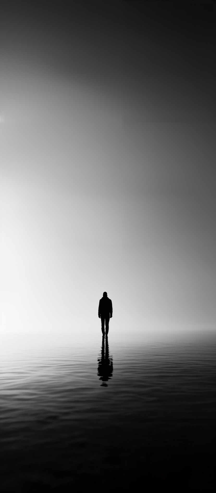 The image is in black and white, showcasing a vast expanse of calm water with a lone figure walking towards the horizon. The figure is silhouetted, making it appear as a dark shape against the lighter background. The water reflects the figure, creating a mirrored image. Above, the sky is vast and clear, transitioning from a darker shade at the top to a lighter one near the horizon, suggesting either dawn or dusk.