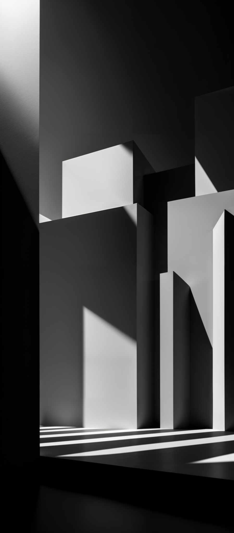 The image showcases a monochromatic scene with varying shades of gray. It features geometric shapes, predominantly rectangular and cubic, that are juxtaposed against each other. The play of light and shadow creates a dramatic effect, highlighting the edges and surfaces of the objects. The composition is symmetrical, with the light source coming from the top left, casting shadows on the right side of the shapes.