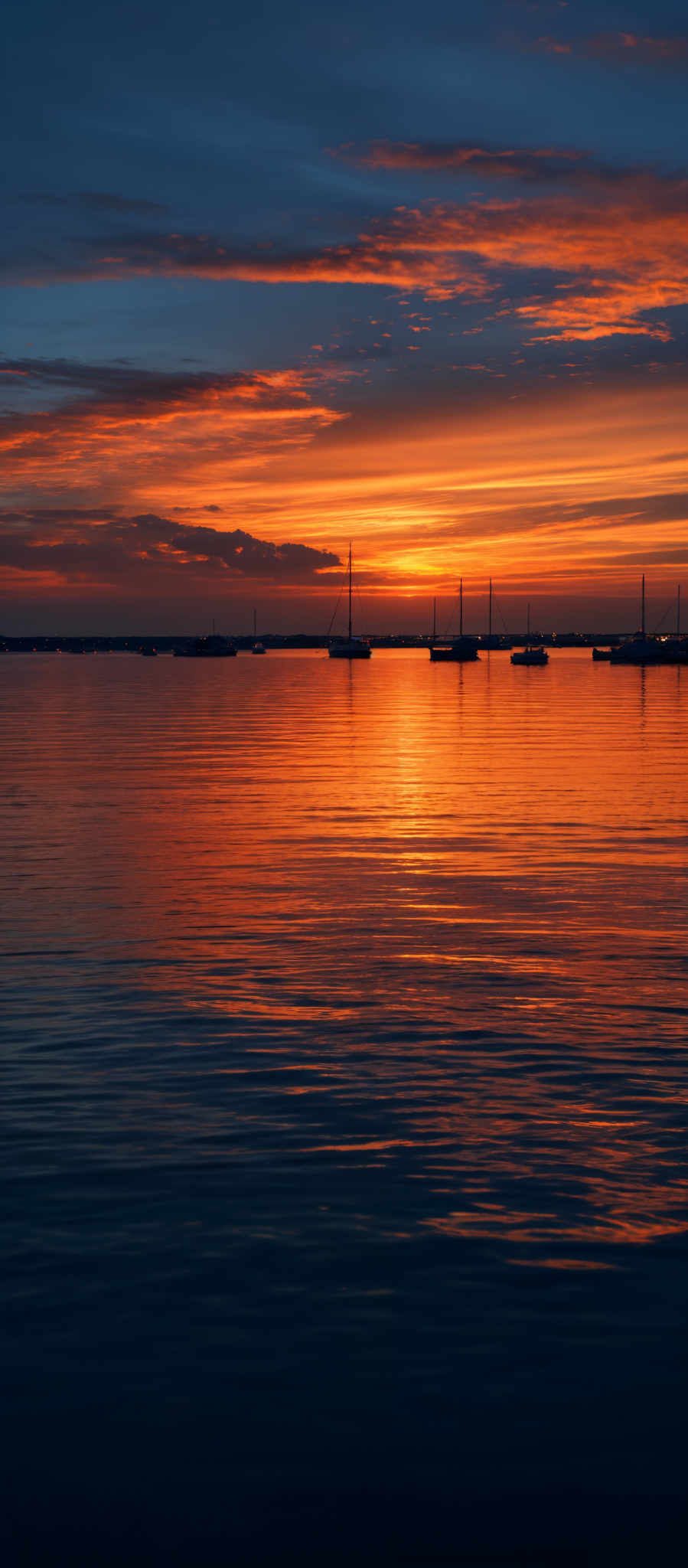 The image showcases a breathtaking sunset over a calm body of water. The sky is painted with hues of orange, gold, and deep blue, with the sun setting near the horizon. The sun's reflection creates a shimmering path on the water's surface. Silhouetted against the vibrant sky are several boats, anchored at a distance, with their masts pointing upwards. The horizon is slightly obscured by a thin layer of clouds, adding depth to the scene.