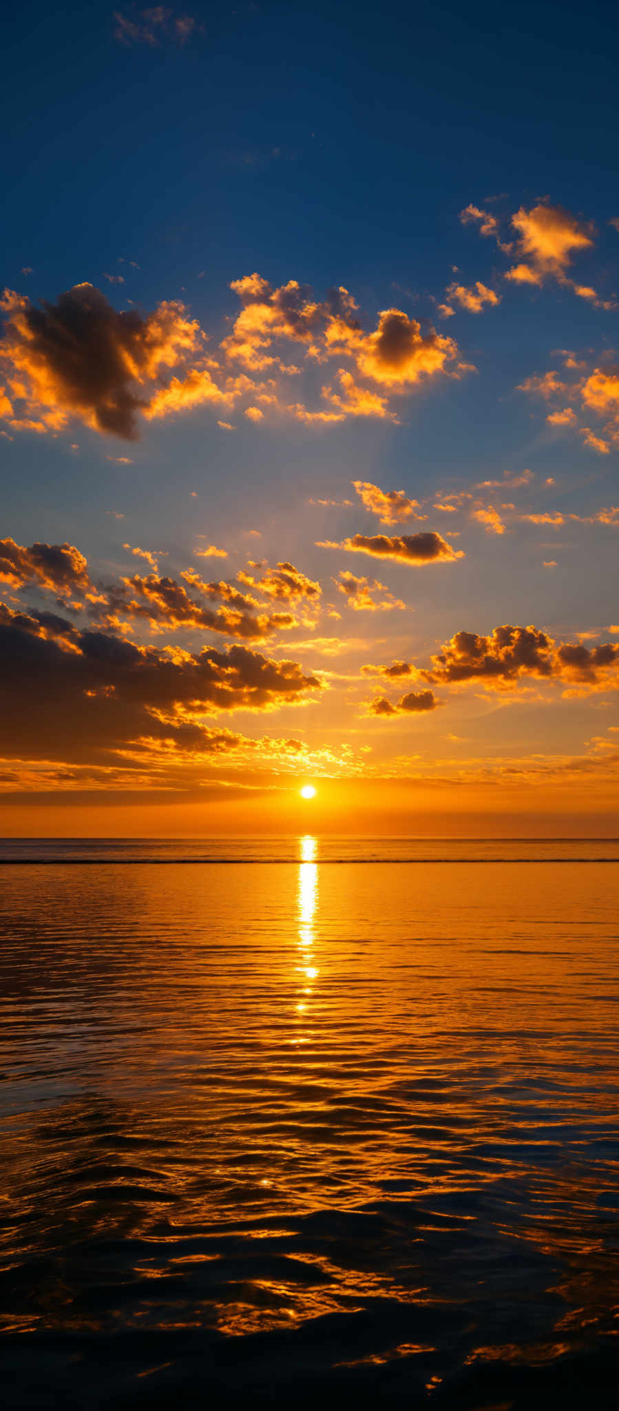 The image showcases a breathtaking sunset over a calm body of water. The sky is painted with hues of blue, transitioning to golden yellow and deep orange near the horizon. The sun is captured just above the waterline, casting a brilliant reflection on the water's surface. The clouds are scattered across the sky, with some appearing fluffy and others taking on a more elongated shape. The overall ambiance of the image is serene and tranquil, evoking feelings of peace and wonder.