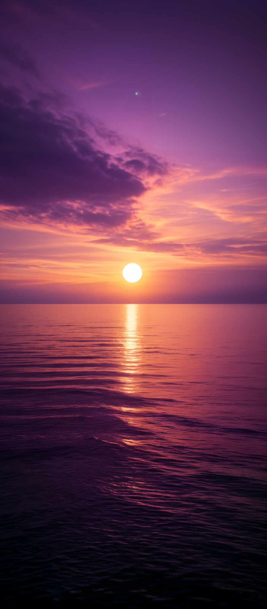 The image showcases a breathtaking sunset over a calm sea. The sky is painted in hues of purple, pink, and orange, with the sun nearing the horizon casting a golden reflection on the water. The sun's reflection creates a serene pathway of light across the sea, with gentle ripples disturbing the surface. Above, the sky is adorned with a few scattered clouds, adding depth and texture to the scene.