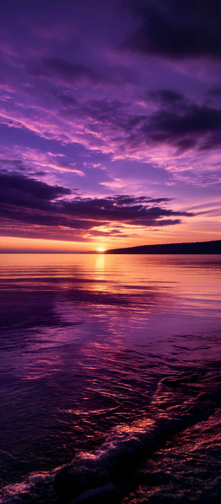 The image showcases a breathtaking sunset over a calm sea. The sky is painted with hues of purple, pink, and orange, with the sun nearing the horizon casting a golden glow. The clouds are scattered, reflecting the colors of the sunset. The sea mirrors the sky's colors, with gentle waves lapping the shore. In the distance, there's a silhouette of a landmass or island.