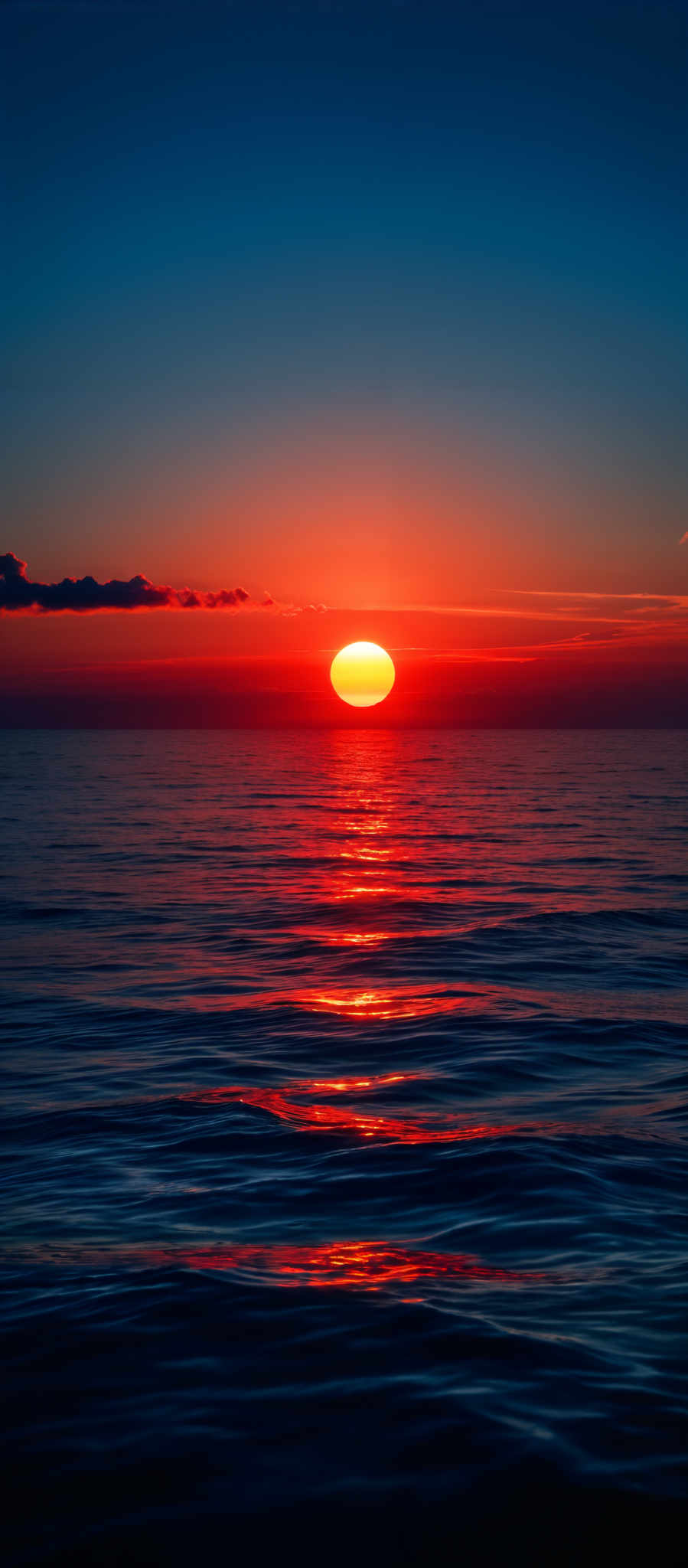 The image showcases a breathtaking sunset over a calm sea. The sun is a brilliant orb, casting a fiery orange and red glow, which reflects upon the water's surface. The sky transitions from a deep blue at the top to a gradient of orange and pink closer to the horizon. A few clouds are visible, adding depth and contrast to the scene. The gentle waves of the sea create a rhythmic pattern, with the sun's reflection creating shimmering pathways across the water.