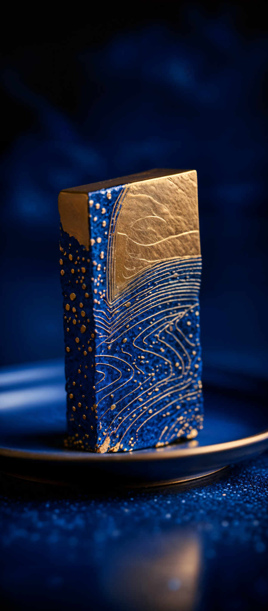 The image showcases a rectangular object with a golden hue on one side and a deep blue on the other. The golden side has intricate patterns resembling waves or ripples, while the blue side has a more textured appearance, possibly mimicking a topographical or geographical map. The object is placed on a plate, and the background is a deep shade of blue, creating a contrast that emphasizes the object's colors.