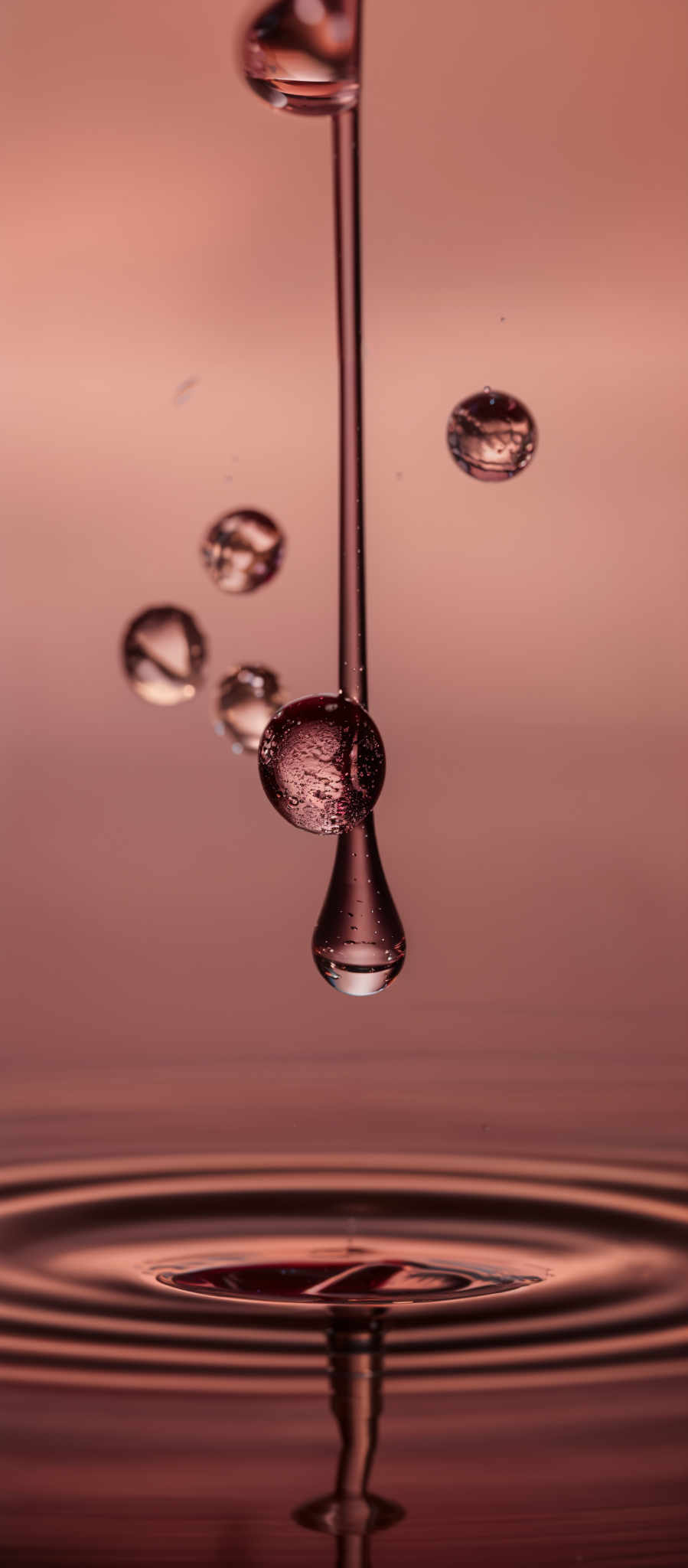 The image showcases a series of water droplets suspended in the air, with one larger droplet at the top appearing to be dripping or falling. The background is a gradient of warm colors, transitioning from a deep red at the bottom to a lighter pinkish hue at the upper part. The water drops reflect light, creating a shimmering effect. The largest droplett at the center has a magnified view, revealing intricate details of its surface, including tiny bubbles and reflections.