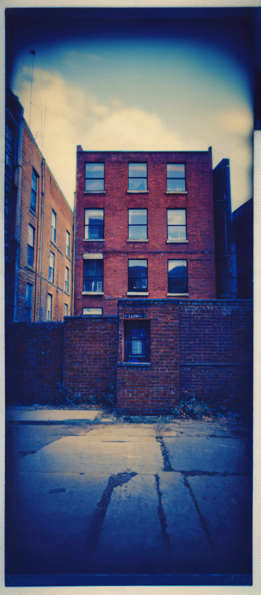 The image showcases a brick building with a rectangular shape. The predominant color of the building is a deep red or brick color. The sky above the building has a gradient of blue, transitioning from a lighter shade at the top to a darker hue towards the horizon. The ground appears to be made of concrete, with visible cracks and patches. There's a small brick structure in the foreground, possibly a utility box or a similar object, with a window. The overall ambiance of the image is somewhat moody, accentuated by the overcast sky and the deep tones of the buildings.