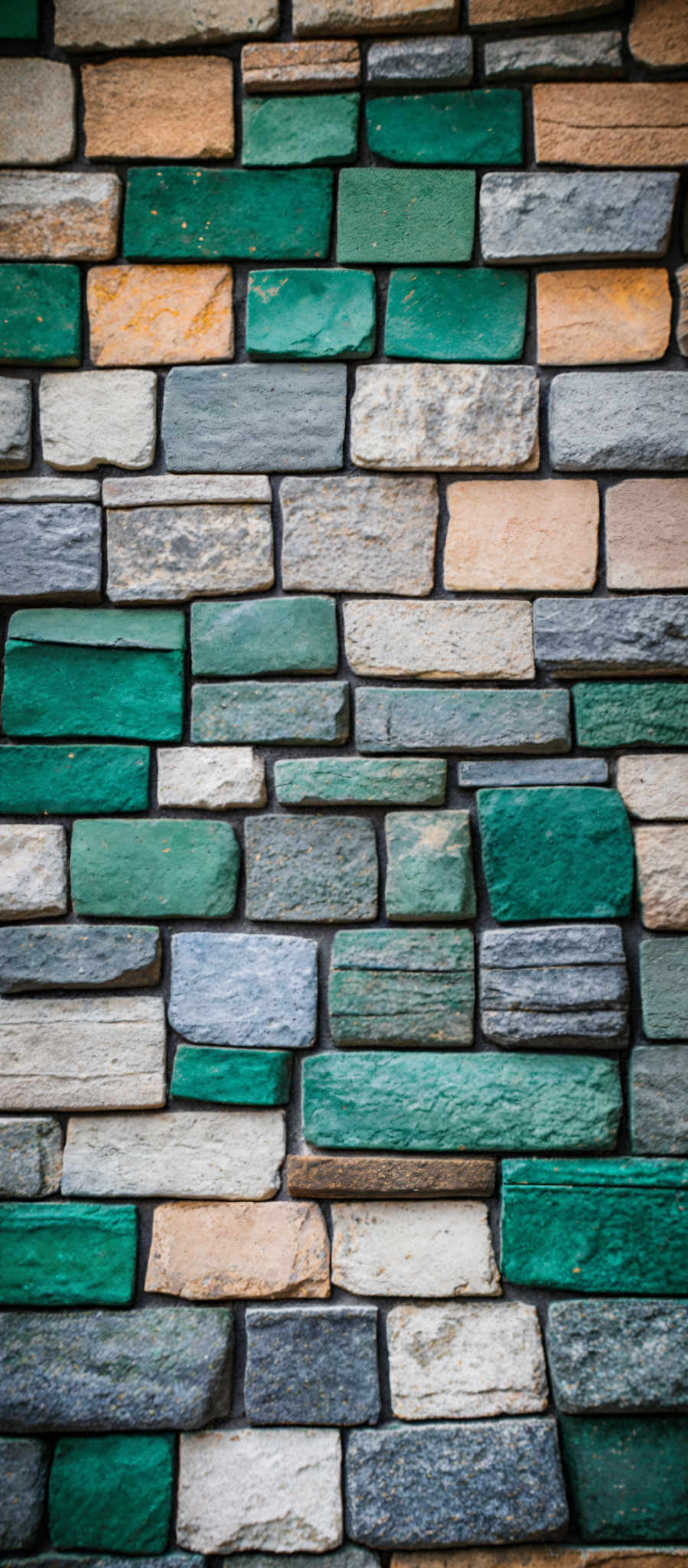 The image showcases a wall made up of rectangular bricks or stones. These bricks vary in color, predominantly in shades of green, beige, and gray. The bricks are of different sizes, with some being narrow and others wider. The texture of the bricks appears to be rough, and there are visible signs of wear and aging on some of them.