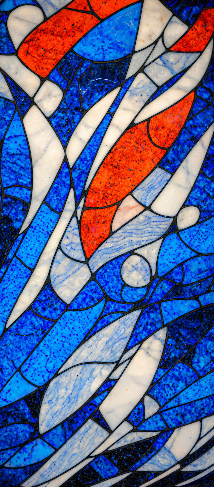 The image showcases a vibrant mosaic pattern. It predominantly features shades of blue, white, and orange. The blue is a deep, rich hue, interspersed with lighter shades, giving it a marbled appearance. The white is used to create contrast, especially in the form of elongated shapes that resemble waves or streamlined objects. The orange is used sporadically, adding a pop of warmth to the cooler tones. The overall design gives an impression of fluidity and movement, reminiscent of water or air currents.