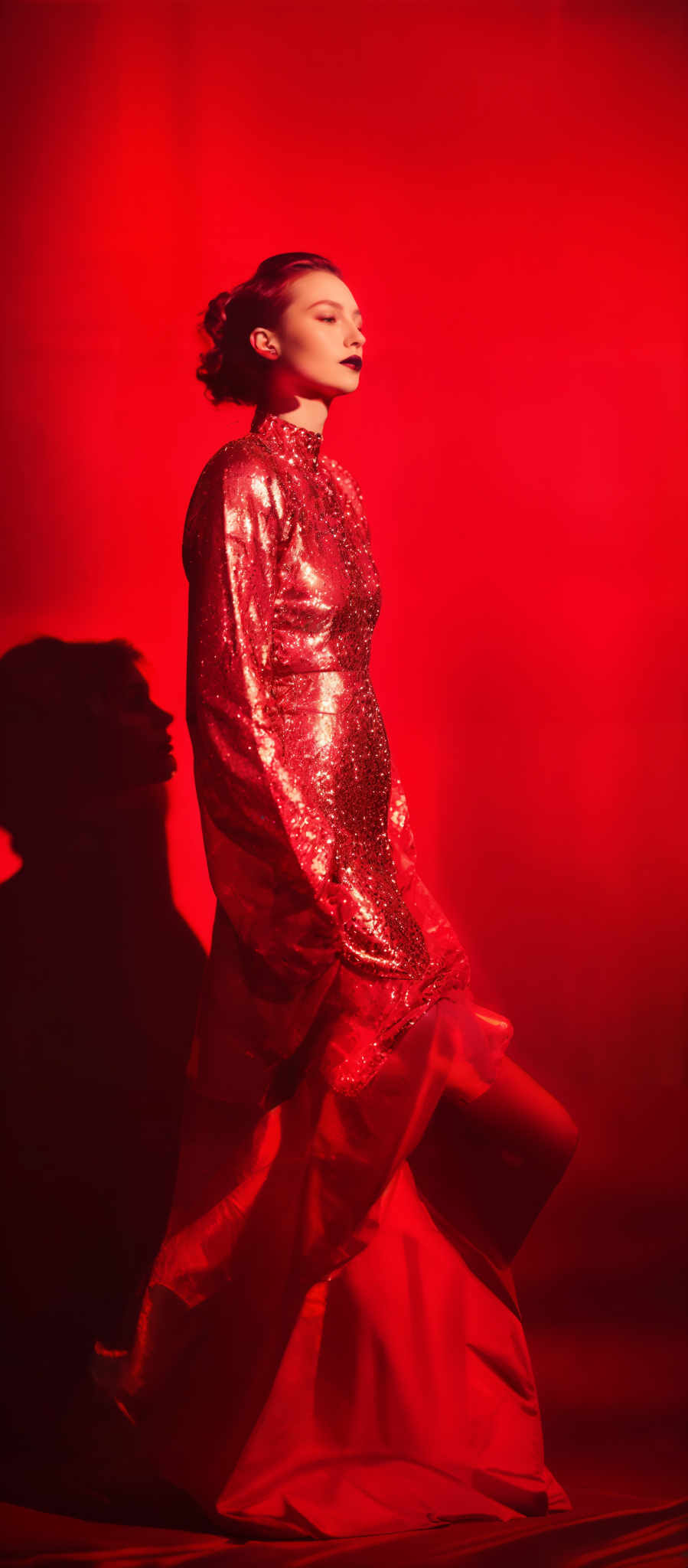 The image predominantly features a rich red hue, creating a dramatic and intense atmosphere. The subject, a woman, is adorned in a shimmering red dress that reflects light, giving it a sparkling appearance. The dress has a high neckline and long sleeves. The woman's makeup is bold, with dark lipstick and a subtle eye makeup. Her hair is styled in an updo, and she appears to be in a contemplative or serene state. The background is mostly dark, with a shadow of the woman' s profile visible, adding depth to the image and emphasizing the subject.