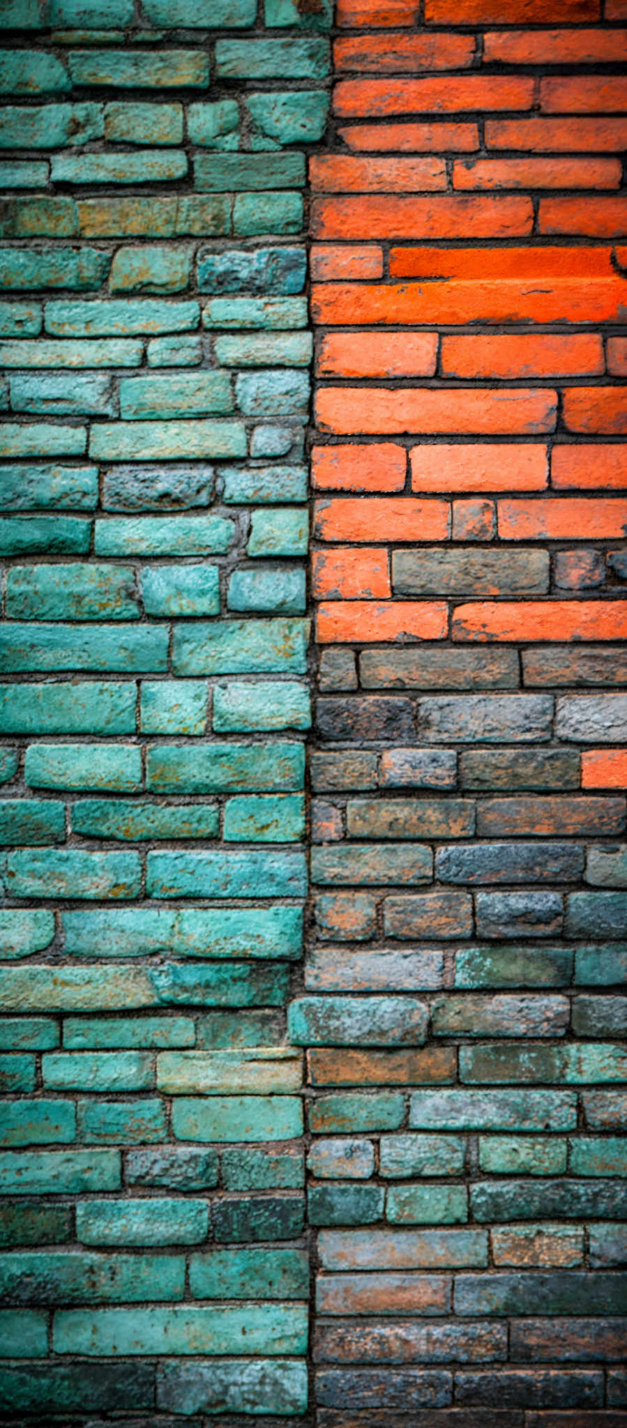 The image showcases a wall made of bricks. The bricks are arranged in a vertical pattern. The colors of the bricks vary from a deep teal to a vibrant orange. The texture of the brickwork appears rough, with some bricks showing signs of wear and aging. The overall appearance is rustic and gives a sense of age and history.