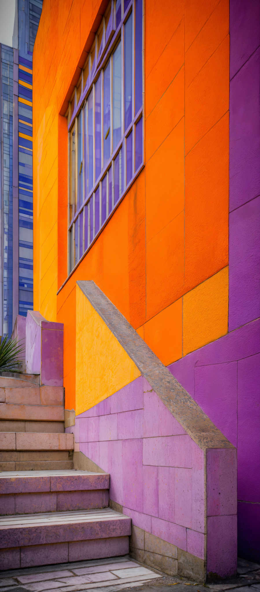 The image showcases a vibrant architectural scene. The dominant colors are orange, purple, and a hint of blue. The structure has a modern design with large windows, some of which are tinted in blue. There's a staircase with steps made of stone, leading up to the entrance of the building. The steps are bordered by a combination of stone and purple tiles. The walls of the structure are made of large square tiles, with the orange wall being the most prominent. The overall design gives a contemporary and artistic vibe.