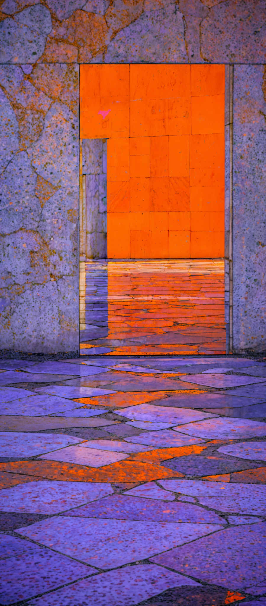 The image showcases a vibrant and colorful architectural setting. The dominant colors are shades of blue, purple, and orange. The architecture consists of large stone or marble blocks, with some showing signs of wear and age. There's a large rectangular opening or doorway in the center, framed by a stone wall. Through this opening, a bright orange wall is visible, reflecting the surrounding environment. The floor is made up of geometric tiles, alternating between blue and purple shades, with reflections of the orange wall and the sky.