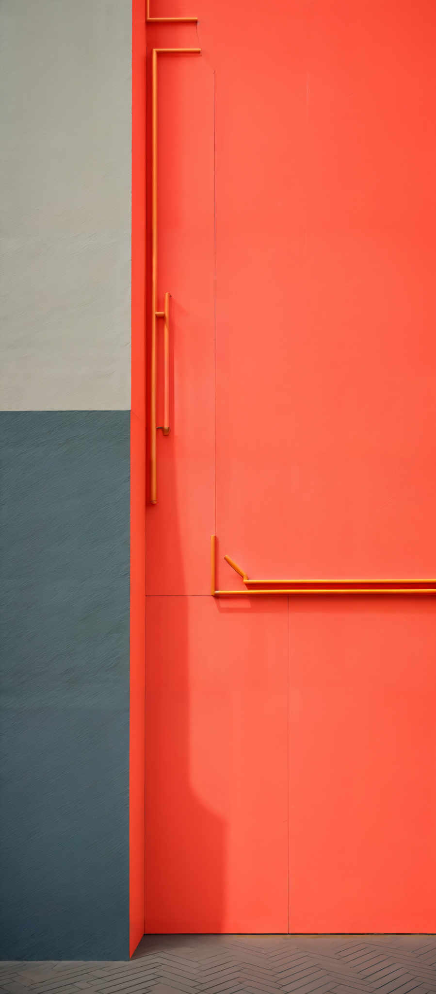 The image showcases a geometric composition of three distinct colors. On the left, there's a gray vertical section. Adjacent to it, on the right, is a vibrant orange wall with a few yellow pipes protruding from it. Below the orange wall, there is a gray horizontal section. The floor appears to be made of gray tiles arranged in a herringbone pattern.