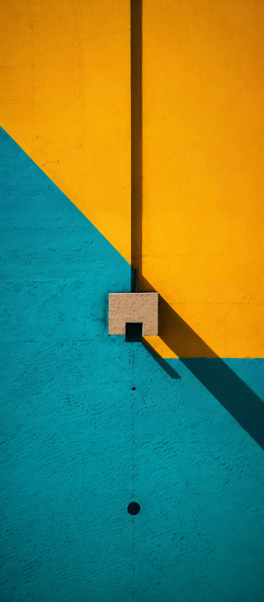 The image showcases a vivid contrast of colors. The upper half is dominated by a bright yellow, while the lower half is a deep turquoise. In the middle, there's a vertical structure, possibly a beam or a pillar, that casts a sharp shadow on the yellow wall. The shadow is elongated and appears to be slightly distorted, possibly due to the angle of the sun or the camera's perspective. The texture of the walls seems rough, with visible brush strokes and imperfections. There's also a small circular object, possibly an air vent or a drain, on the turquose wall.