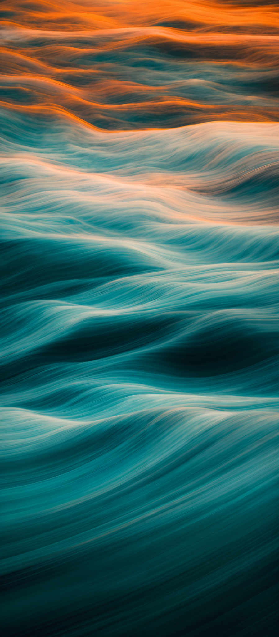 The image showcases a vibrant and dynamic scene of flowing water. The water appears to be in motion, creating a smooth, wavy texture. The color palette is dominated by shades of blue, which gives the water a serene and deep appearance. Above the water, there's a contrasting burst of colors, primarily orange and yellow, which might represent the sunset or sunrise. The overall effect is a blend of calmness from the water and energy from the colors above.