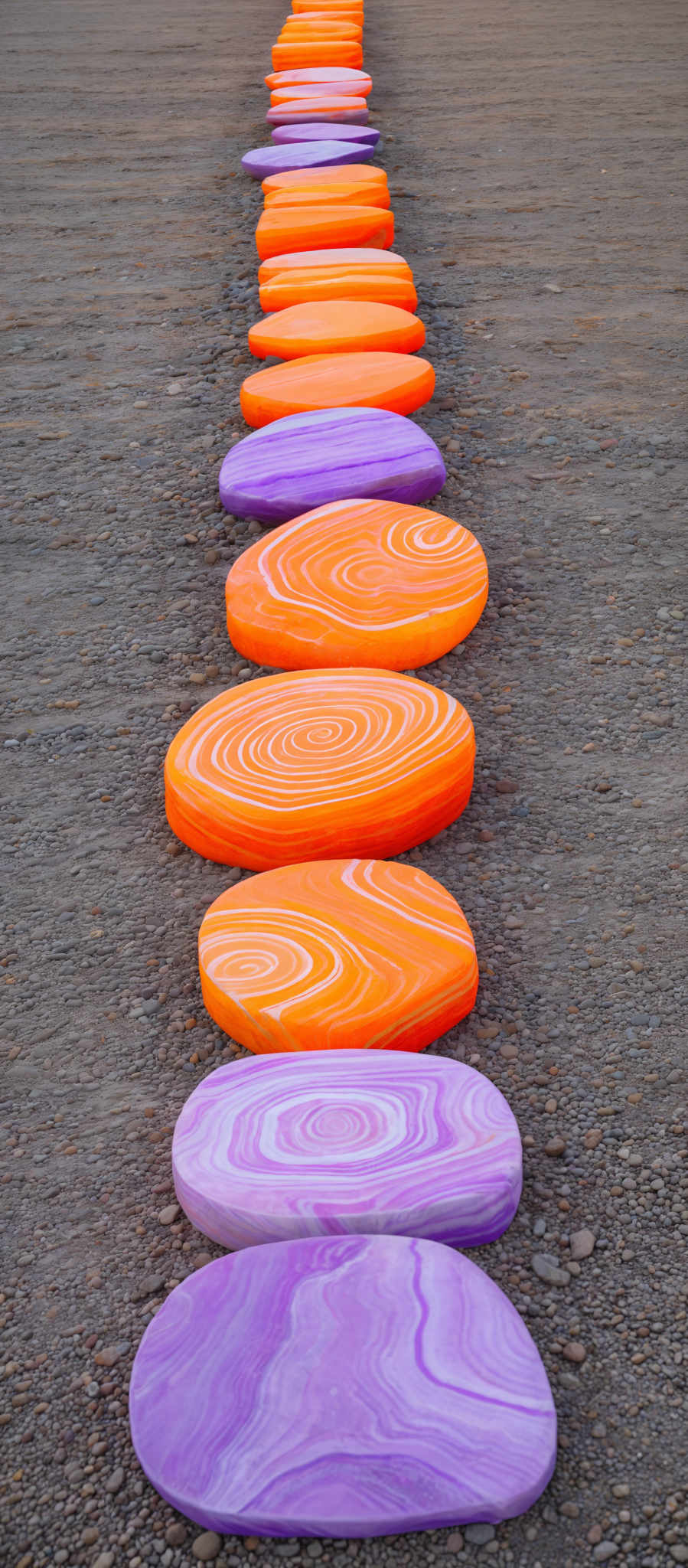 The image showcases a series of oval-shaped objects arranged in a line on a gravelly ground. These objects are vibrant in color, with hues of orange, purple, and white. The orange objects have swirling patterns, while the purple ones have a more uniform, wavy design. The white patterns on the orange objects resemble concentric circles.