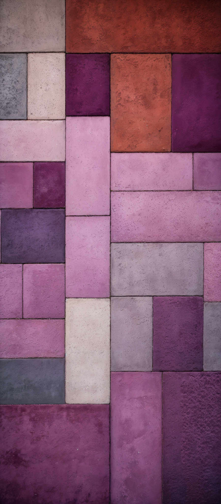 The image showcases a pattern of rectangular and square tiles arranged in a grid format. The tiles come in a variety of colors including shades of pink, purple, orange, and gray. The colors are not uniform, with each tile having its own unique hue and texture. The shapes range from larger rectangles to smaller squares, and the overall appearance gives a sense of a textured wall or floor.