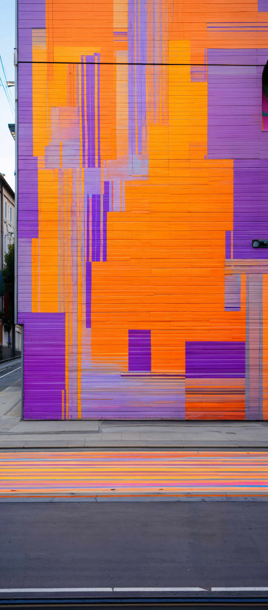 The image showcases a vibrant mural on a building's side. The mural predominantly features shades of orange, purple, and white. The design is abstract, with overlapping geometric shapes and patterns. The colors are bold and contrast sharply with the surrounding environment, which includes a street with colorful car tire streaks and a few buildings in the background.