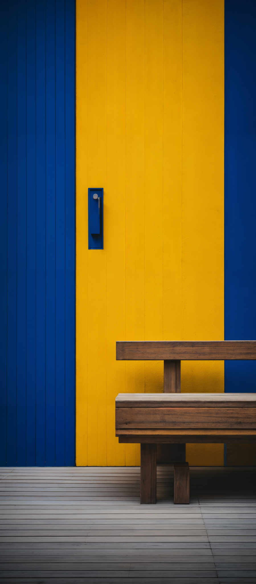 The image showcases a vibrant contrast of colors. On the left side, there's a deep blue wall with vertical wooden panels. On this wall, there is a rectangular, blue-colored mailbox. Adjacent to the blue wall, on the right side, is a bright yellow wall with horizontal wooden panelling. Below the yellow wall, a wooden bench is placed on a wooden floor. The bench has a simple design with two horizontal wooden slats for seating and two vertical wooden supports.