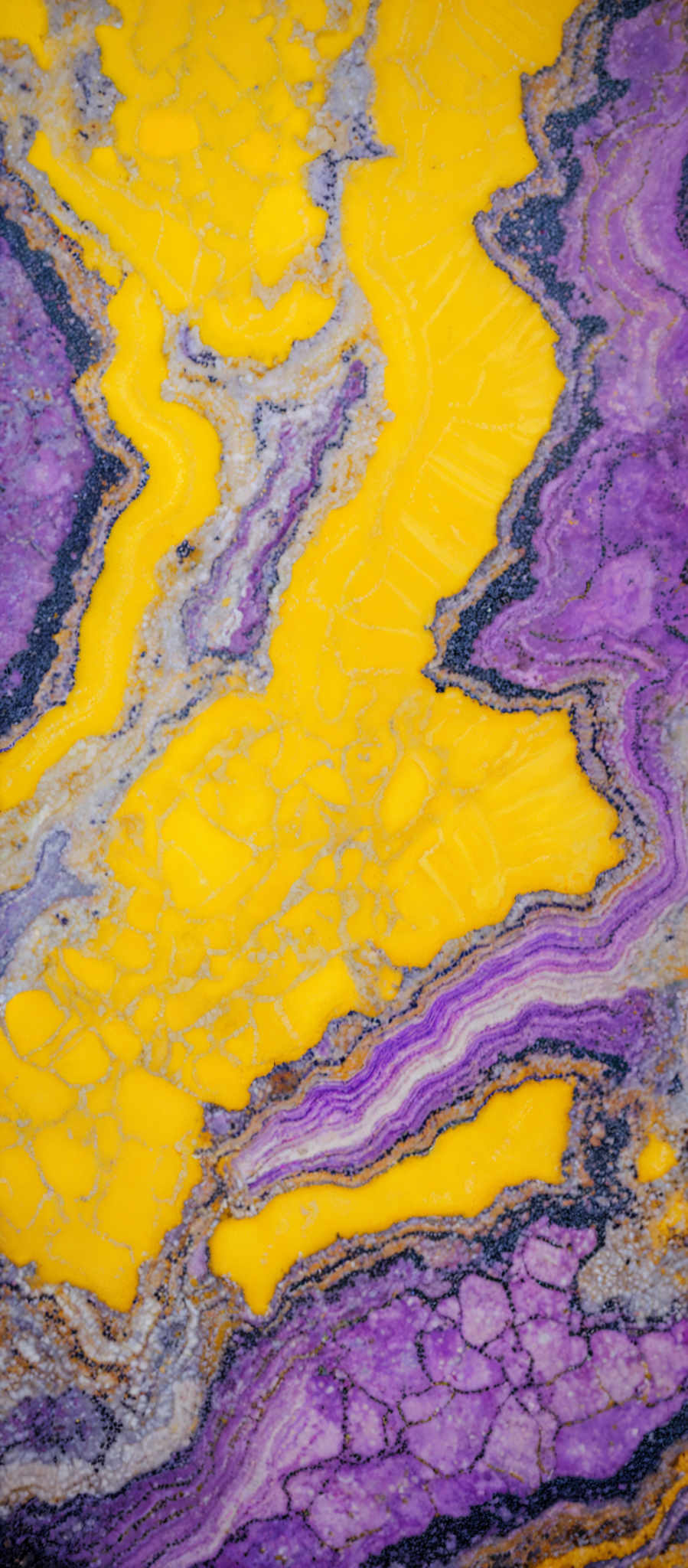 The image showcases a vibrant and intricate pattern of colors. The dominant colors are yellow and purple, with swirls and intricacies that resemble marble or fluid art. The yellow forms wavy patterns, while the purple appears in patches and lace-like designs. There are also areas of gray and white, which add contrast and depth to the overall design.