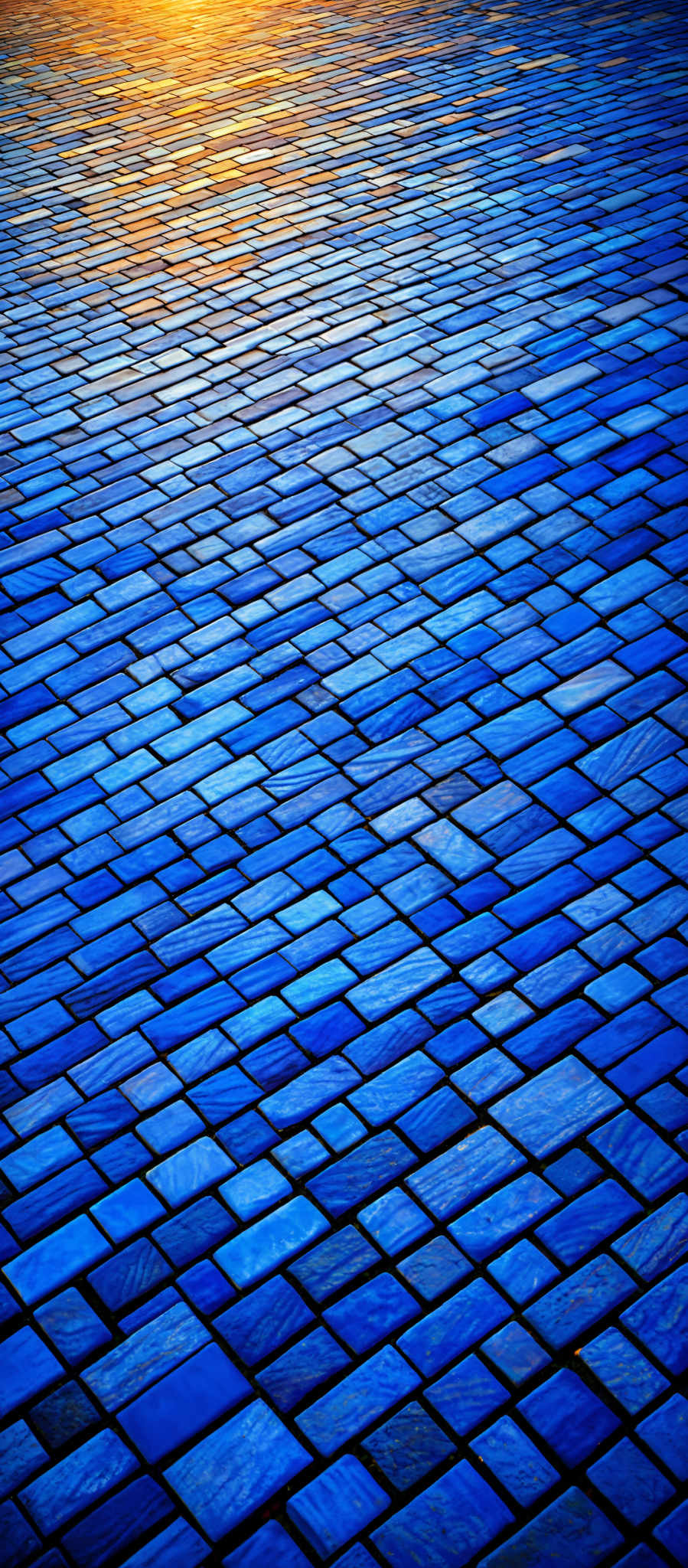 The image showcases a pattern of square tiles arranged in a rhythmic manner. The tiles are predominantly blue with varying shades, ranging from light to dark. The pattern creates a sense of depth and perspective, leading the viewer's eye towards the horizon where a golden hue, possibly from the setting or rising sun, is visible.