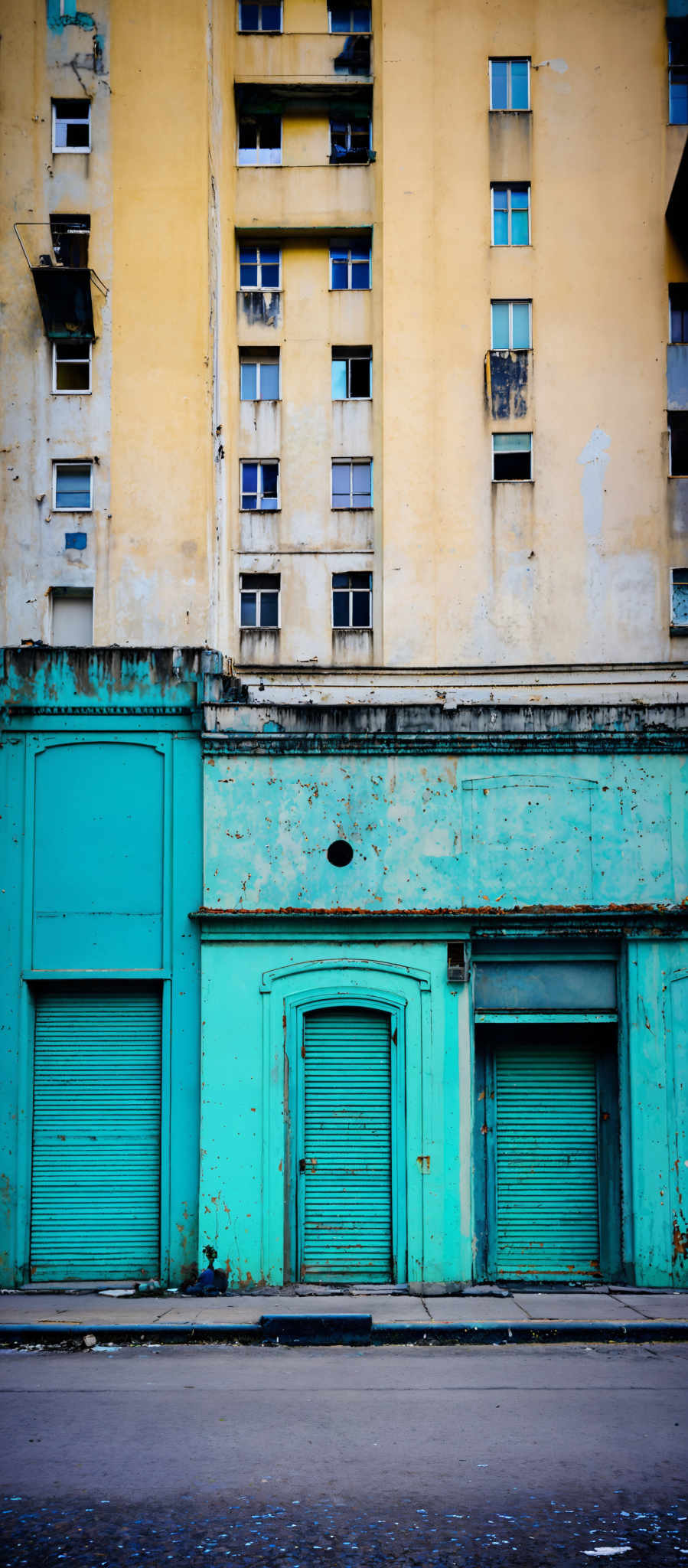 The image showcases a tall building with a pale yellow facade. The building has multiple windows, some of which are open, revealing balconies. The lower part of the image displays a street with three closed teal-colored shops or establishments. Each shop has a single door, and the shutters are rolled down. The street appears to be relatively empty with a few scattered pieces of trash.