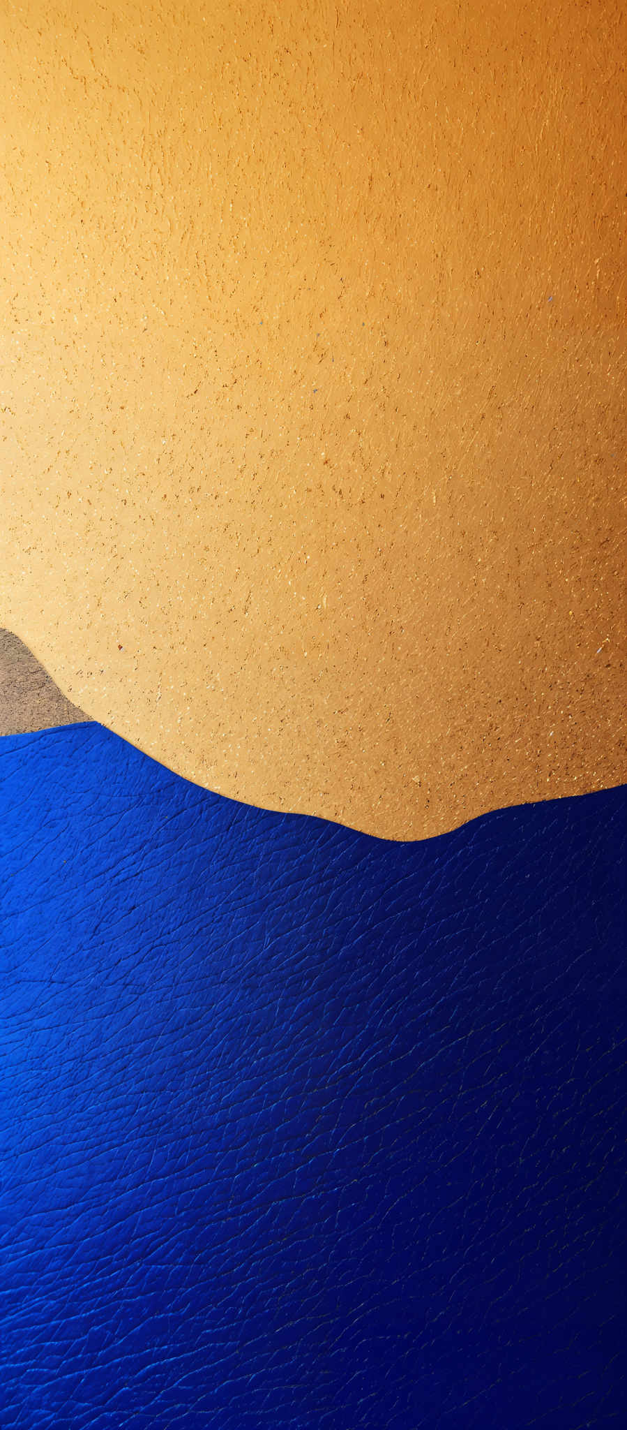 The image showcases a stark contrast between two primary colors: a deep blue and a golden-yellow. The blue represents a vast expanse, possibly a body of water, with a textured surface that suggests ripples or waves. The golden- yellow, on the other hand, appears to depict a sandy beach or coastline. The top portion of the image has a lighter shade of yellow, which gradually transitions into a deeper hue as it moves downwards, possibly indicating the shifting of sand or the reflection of sunlight.