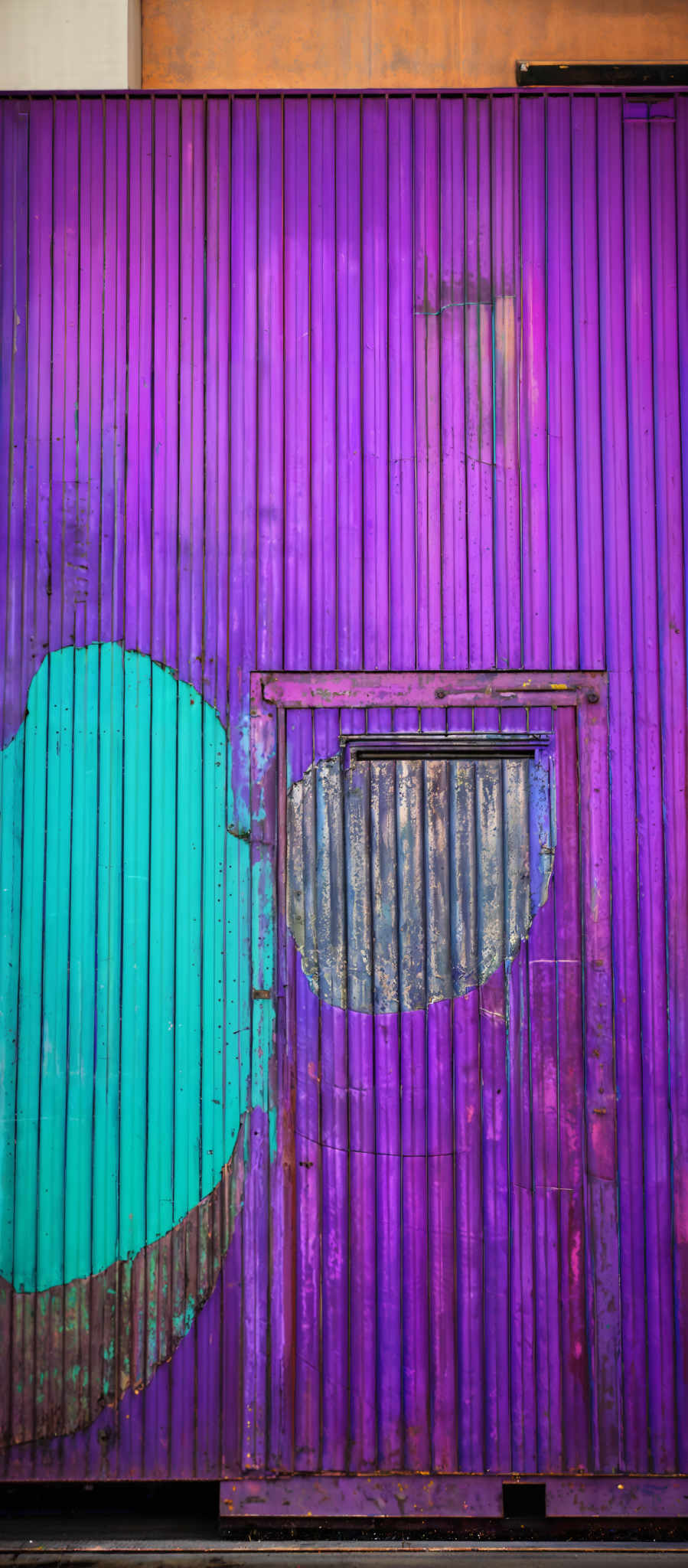 The image showcases a vibrant and textured wall made of vertical wooden planks. The dominant colors are shades of purple and turquoise. There's a large turquoiise shape, resembling a cloud or an abstract form, placed on the wall. Adjacent to it, there's another turquoisie patch with a rusty, weathered appearance. The wooden plates exhibit signs of wear, with some areas showing peeling paint, rust, and other imperfections.