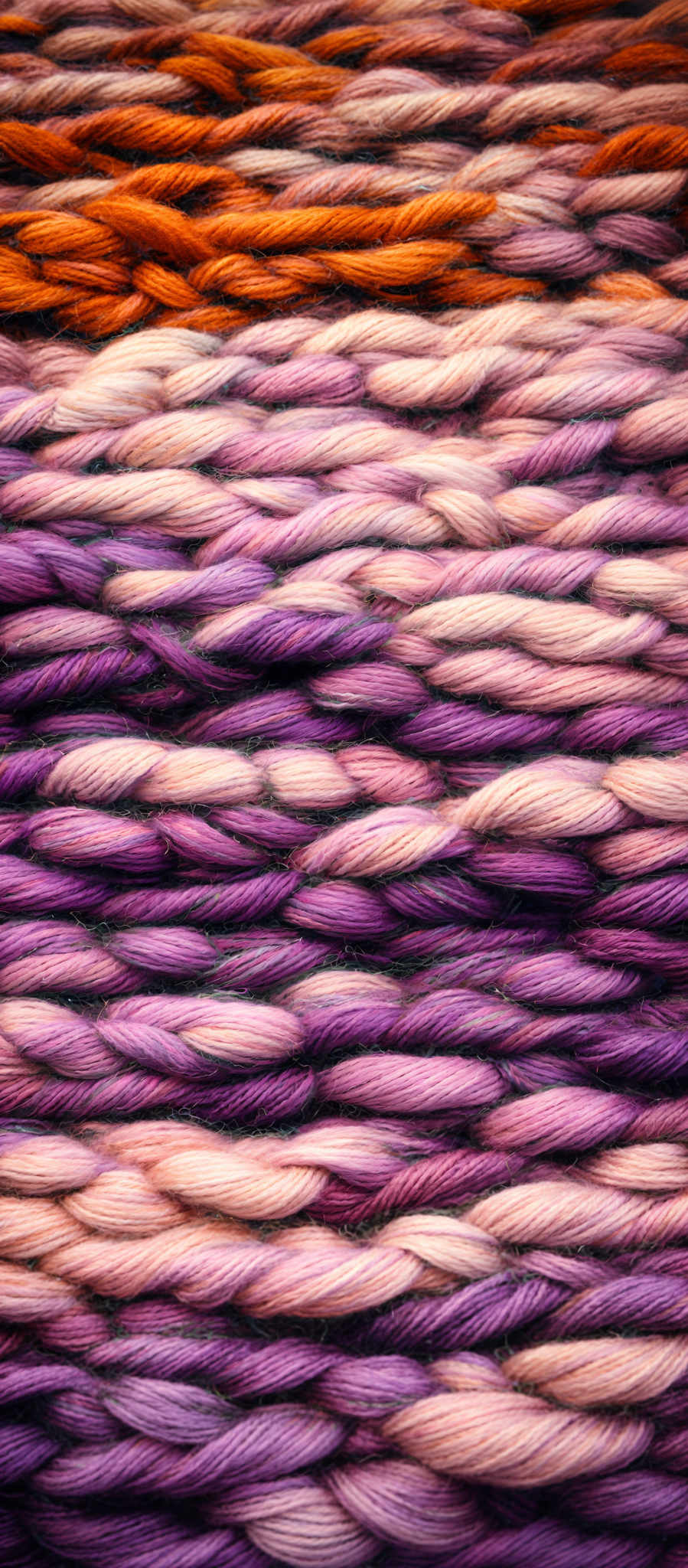 The image showcases a close-up of multiple strands of yarn or thread. These strands are intertwined and layered, displaying a gradient of colors ranging from deep purples and pinks to bright oranges and yellows. The yarn appears to be in the form of braids or twists, with each strand having a twisted or braided shape. The texture of the yarn looks soft and fibrous, and the colors are vibrant, creating a visually appealing and textured pattern.