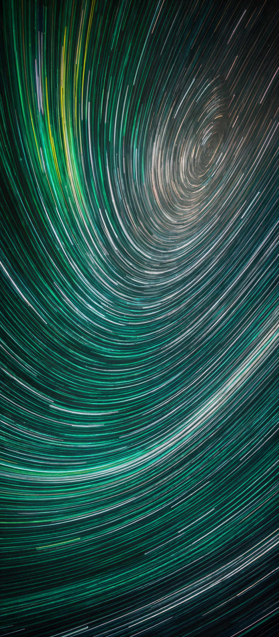 The image showcases a mesmerizing display of star trails, captured over a long period of time. The trails are circular and concentric, with colors ranging from deep blues and greens to bright yellows and whites. The central part of the image has a more intense concentration of trails that appear to spiral inward, creating a captivating vortex effect. The outer edges of the trails gradually fade, giving a sense of depth and dimension to the image, as if the stars are moving inward towards a central point.